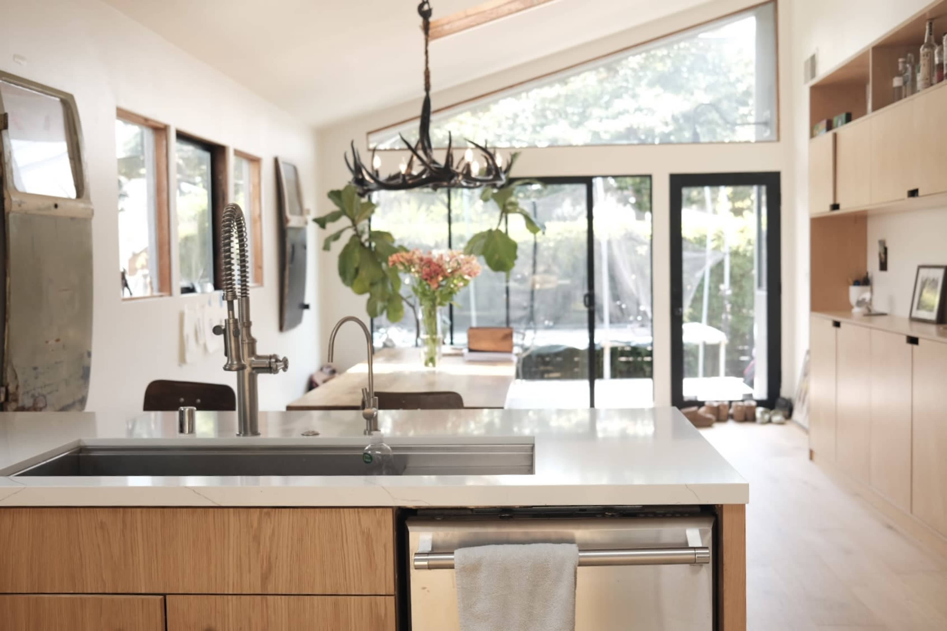 A modern kitchen features a central island with a stainless steel sink, a wooden cabinet, and large windows that provide a view of an outdoor dining space.
