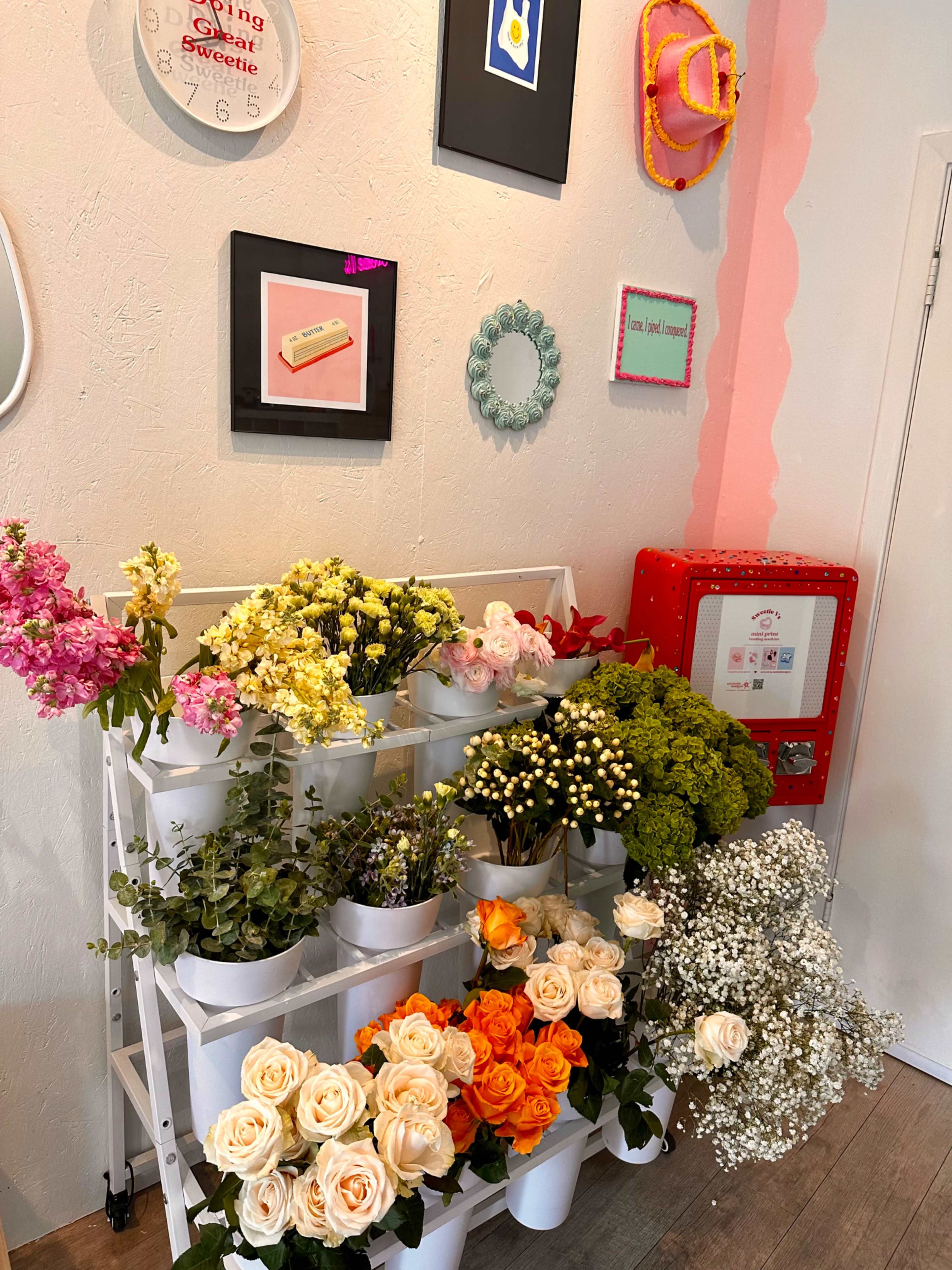 A wall display features various potted flowers in a white shelf, surrounded by framed artwork and decorative items.