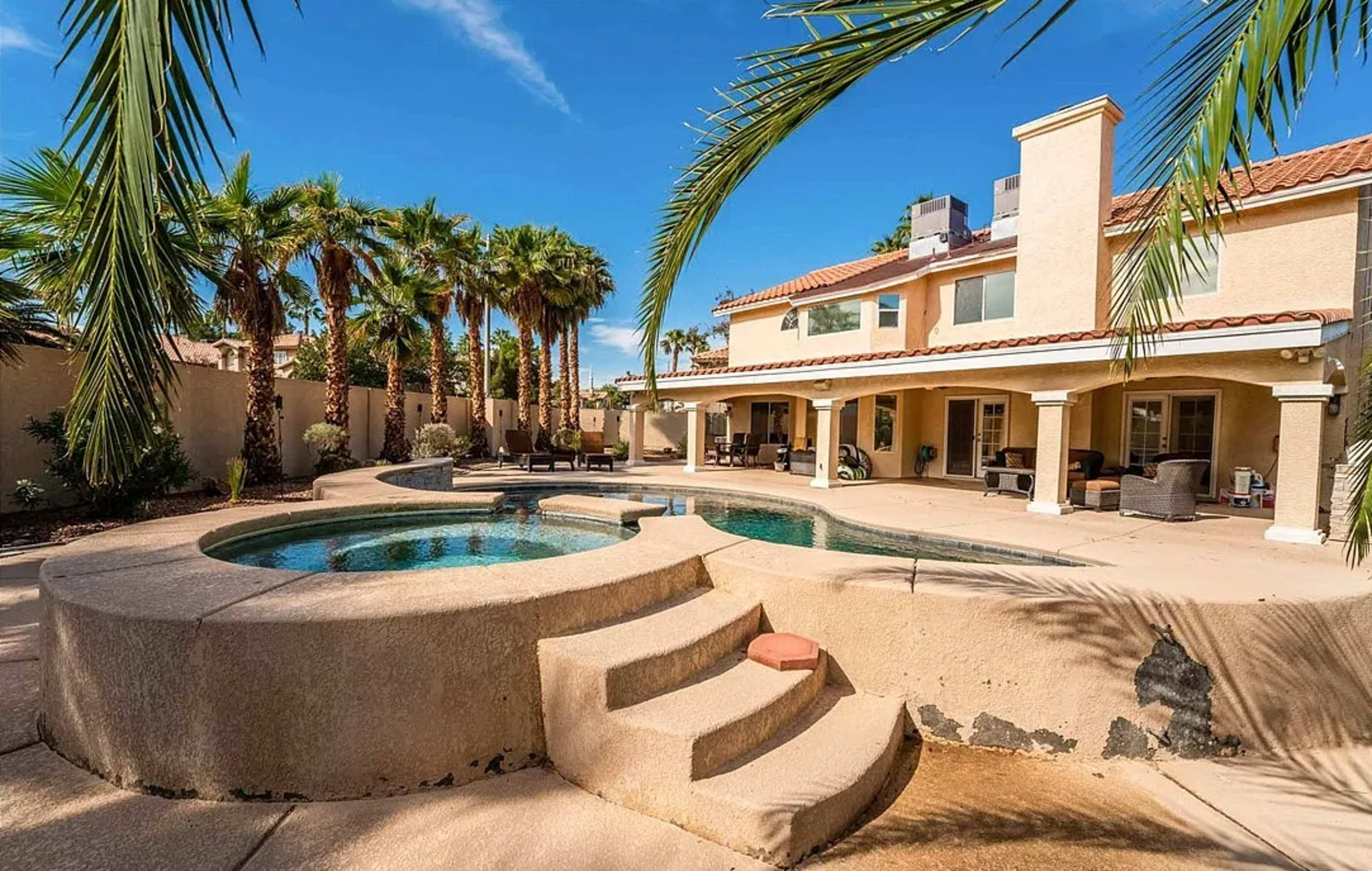 A backyard features a swimming pool with a hot tub, surrounded by tall palm trees and a spacious patio area.