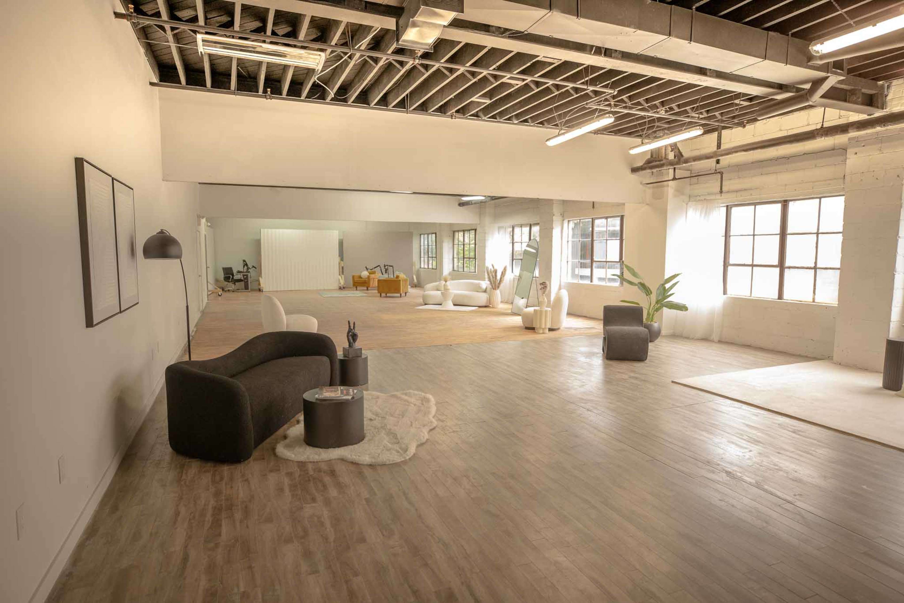 The image shows a spacious, well-lit interior with a seating area and various furniture pieces arranged across a light-colored wooden floor.