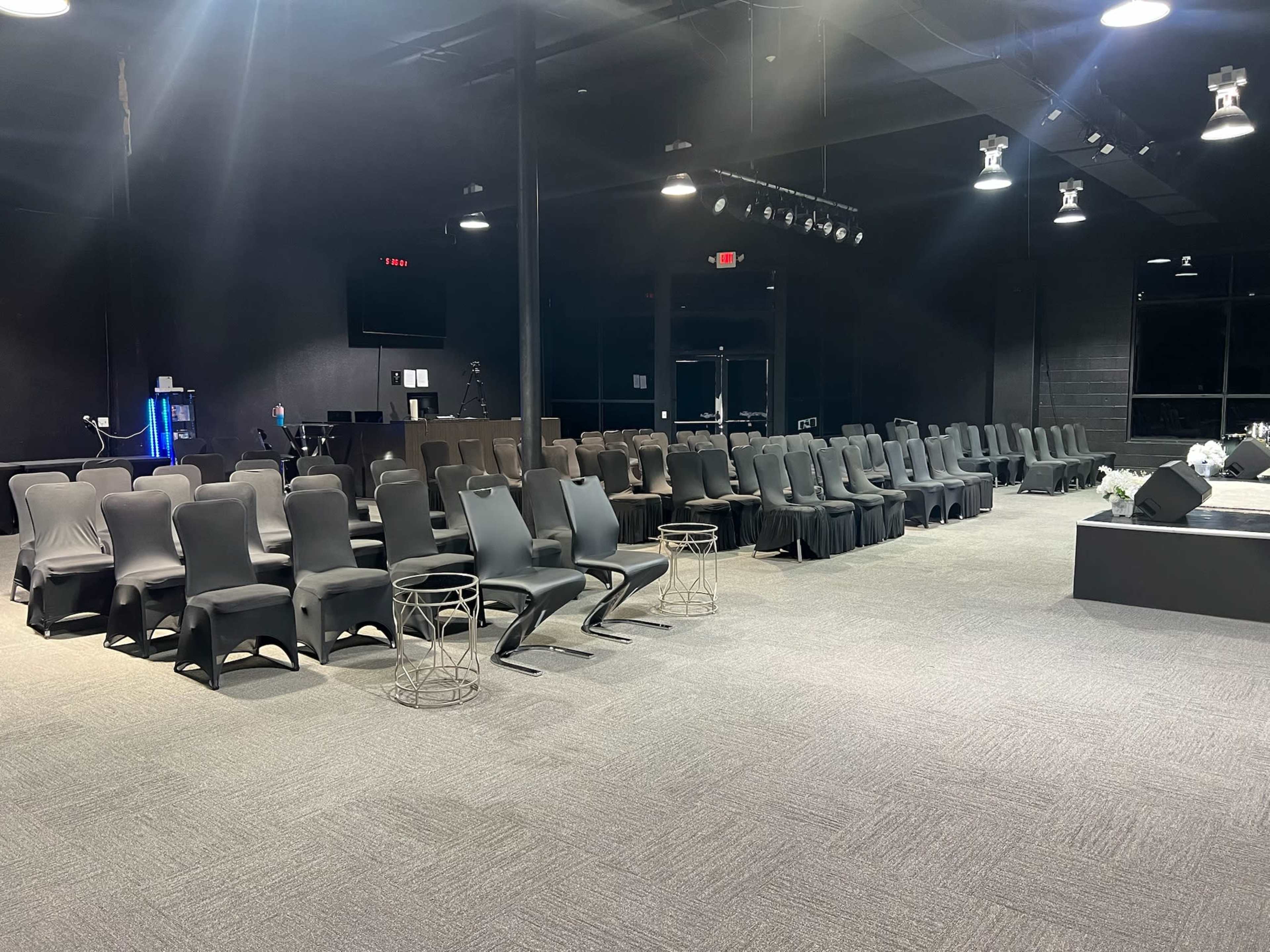 The image shows a darkly lit auditorium with rows of black chairs arranged in a neat formation, accompanied by a stage area.