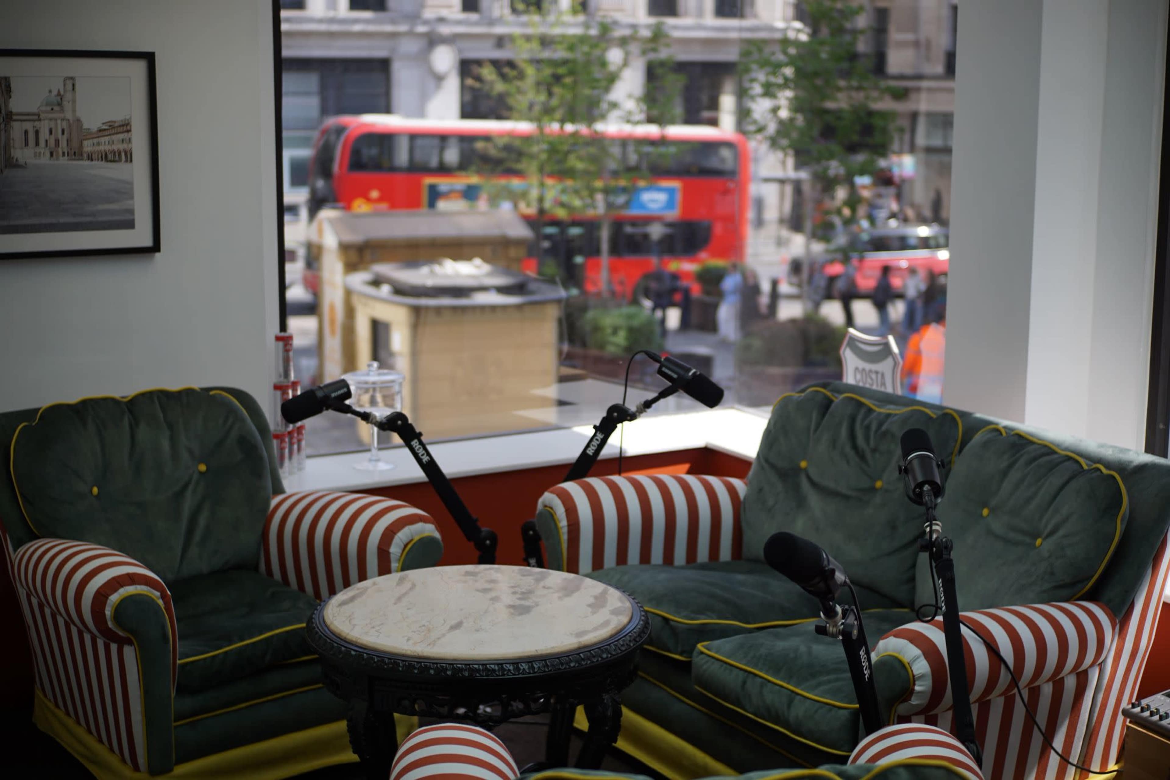 The image shows two striped armchairs facing a round table, with a view of a busy street and a red double-decker bus visible through a large window.