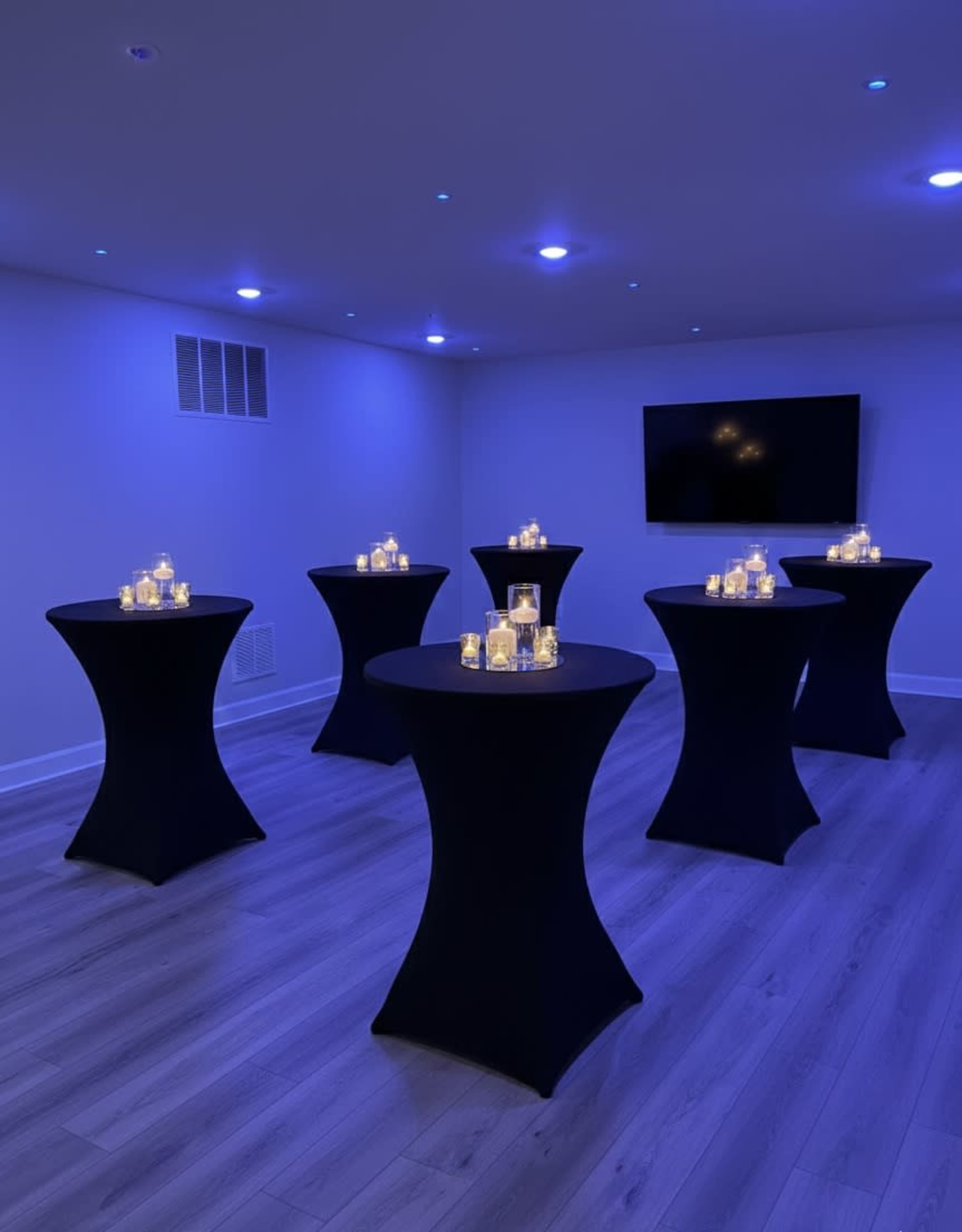 The image shows a dimly lit room with multiple black cocktail tables, each adorned with candles, creating a cozy atmosphere.