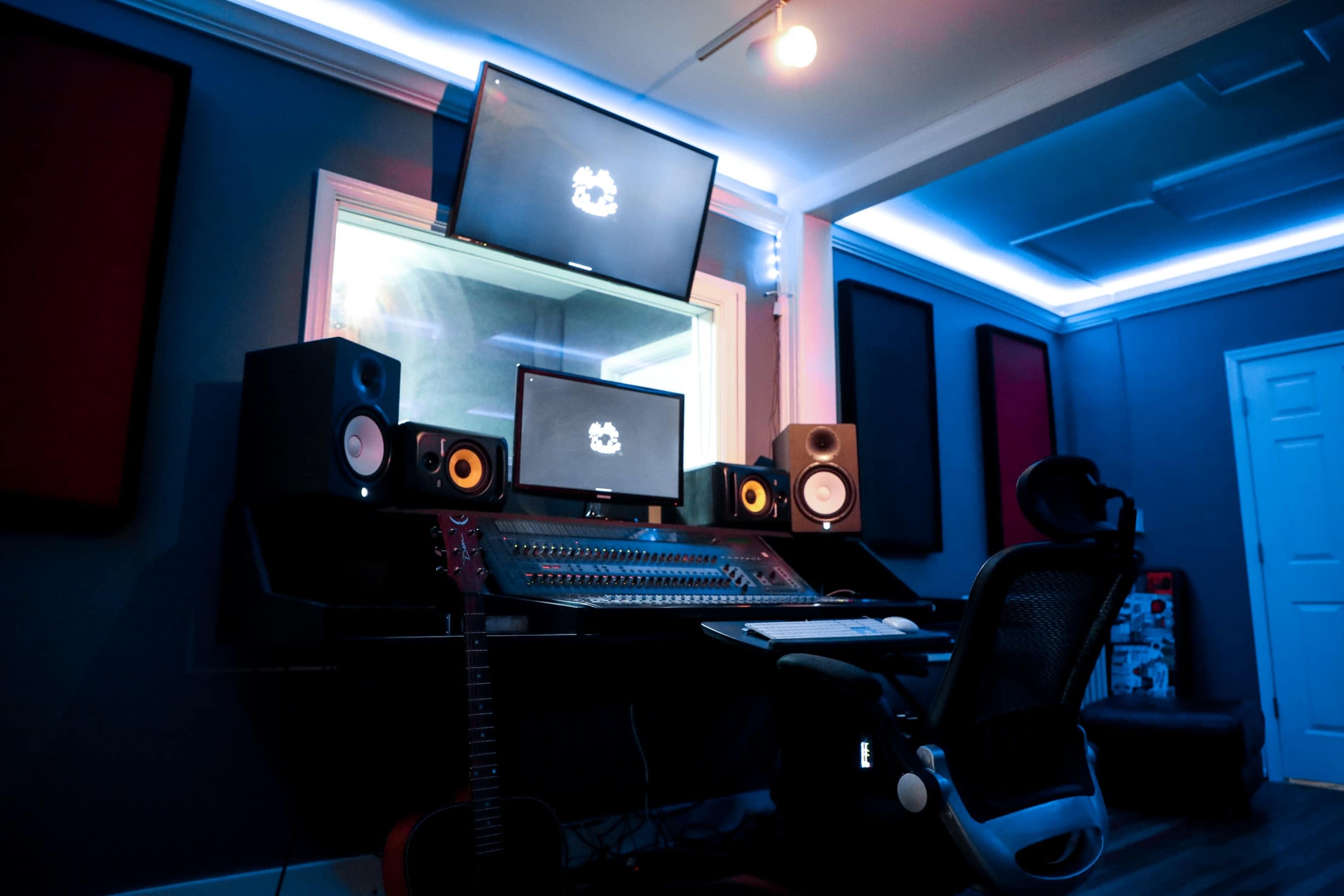 A music studio is equipped with multiple monitors, speakers, a mixing console, and a chair, all illuminated by blue LED lighting.