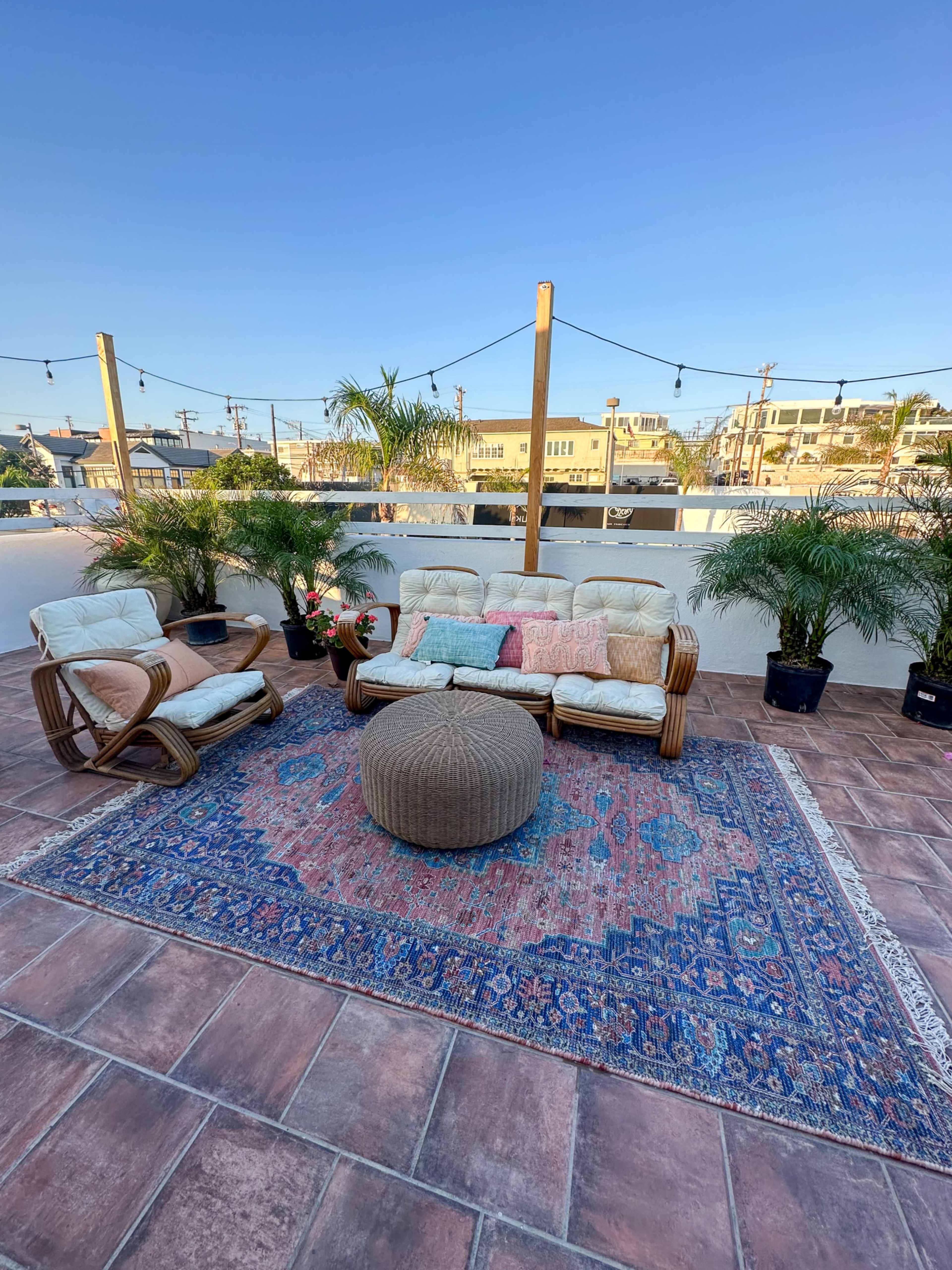 A rooftop seating area features two chairs and a couch on a patterned rug, surrounded by potted plants and with a clear blue sky above.