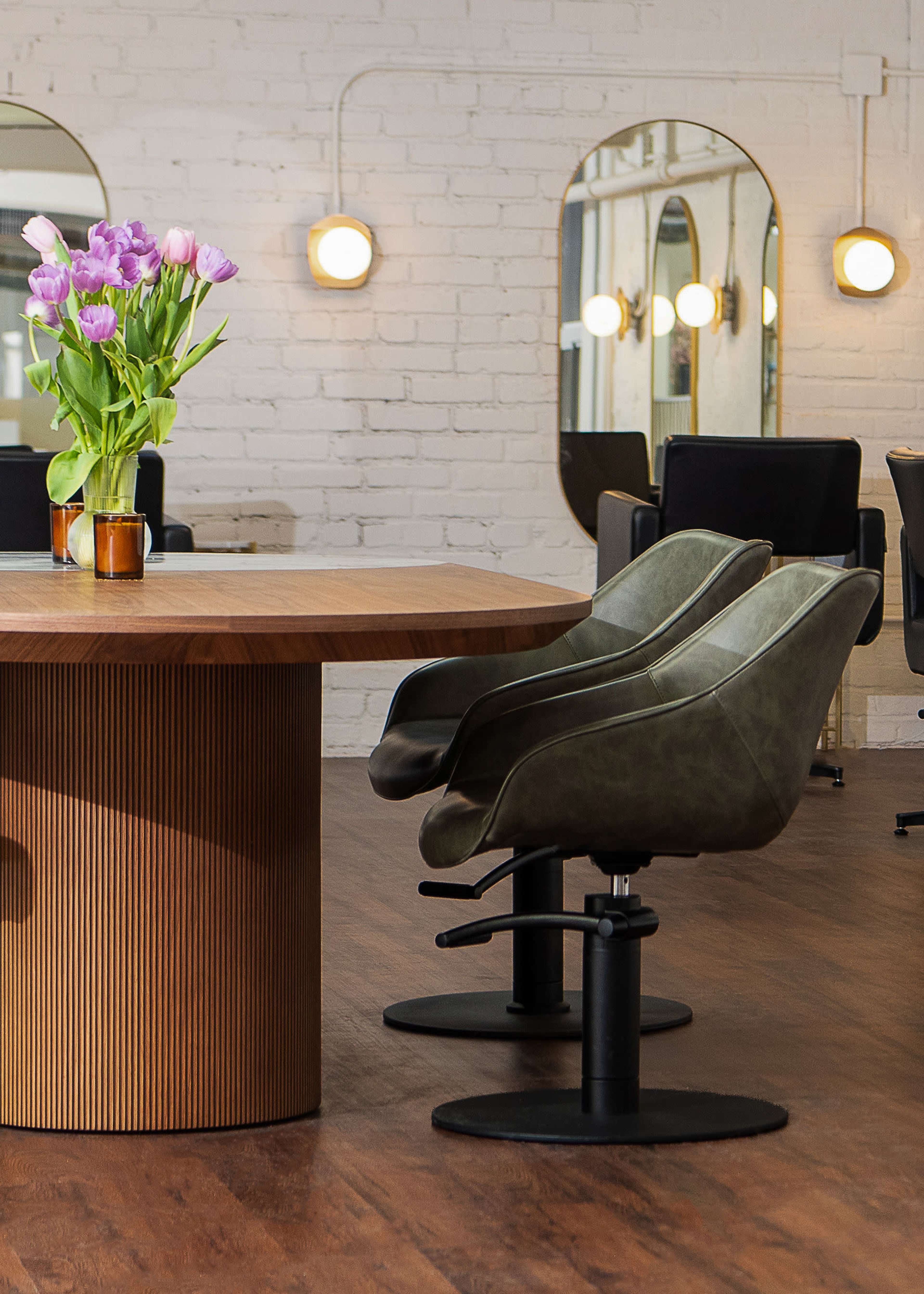 A modern salon interior features a round wooden table, green chairs, and a vase of purple tulips, accented by wall mirrors and soft lighting.