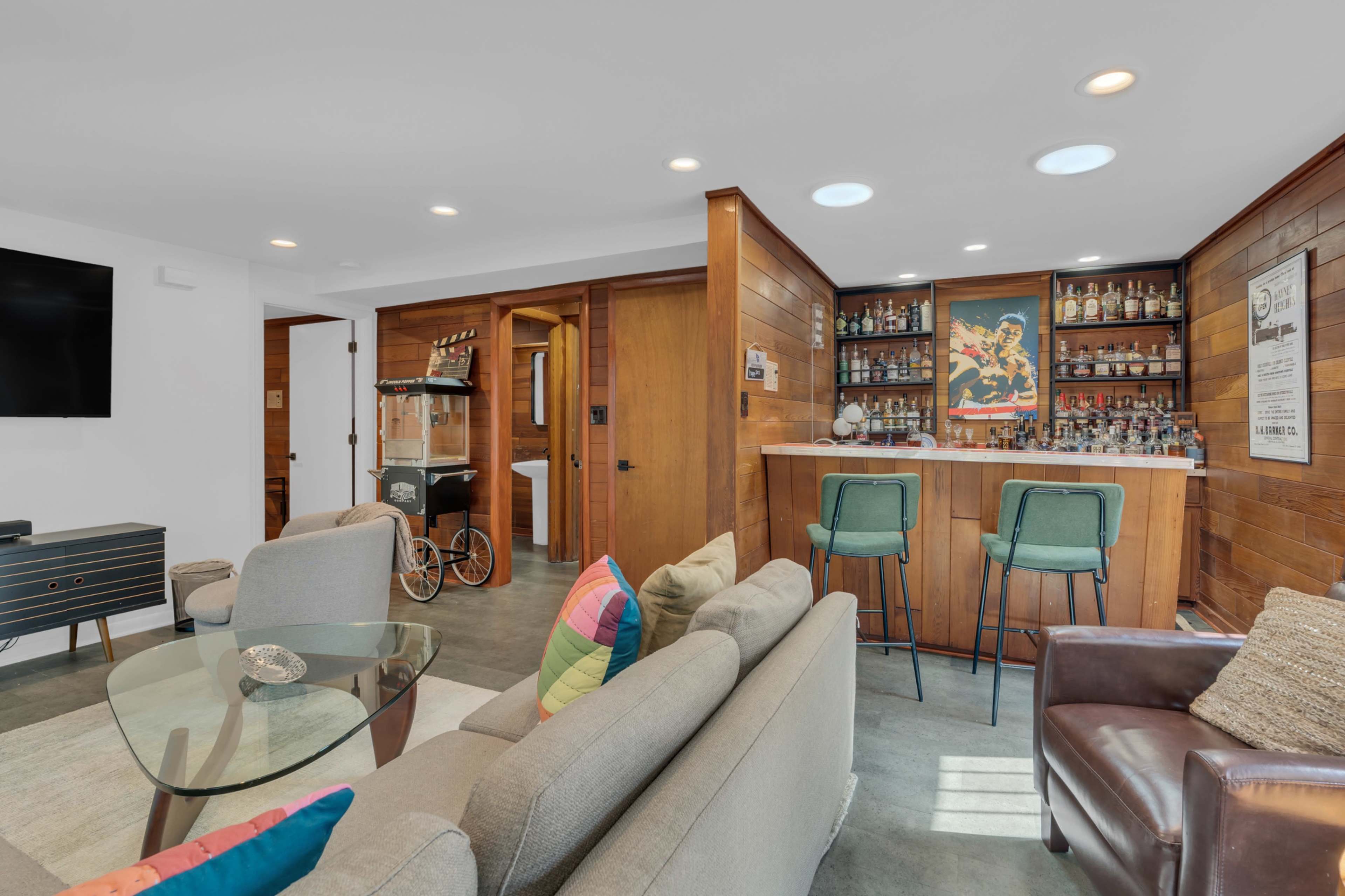 The image shows a modern living space with a bar area, featuring a wooden bar, shelves of bottles, and seating, alongside a cozy sitting area with a coffee table and colorful cushions.
