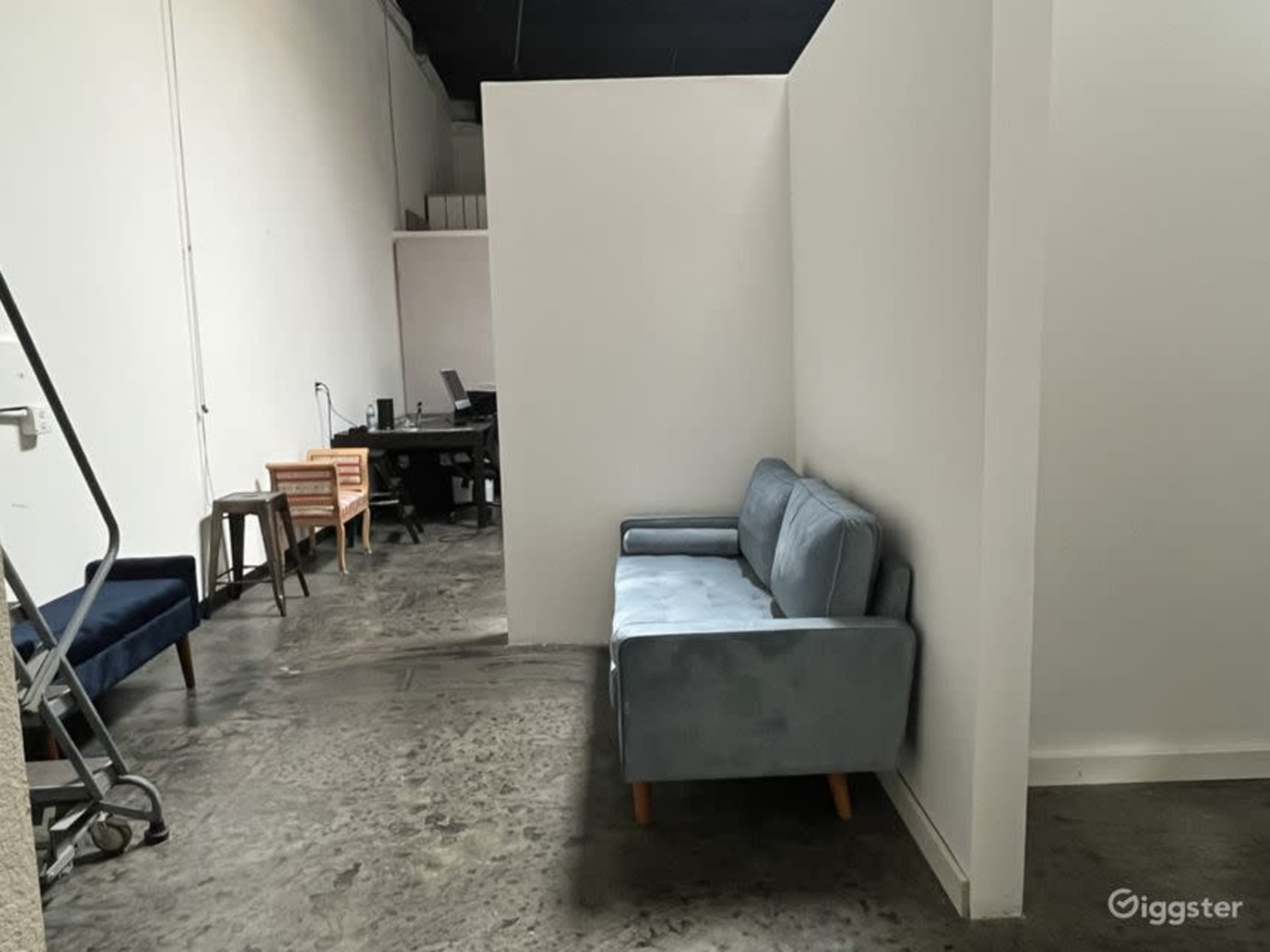 A gray couch positioned against a white wall in an indoor space, with a desk and chair visible in the background.