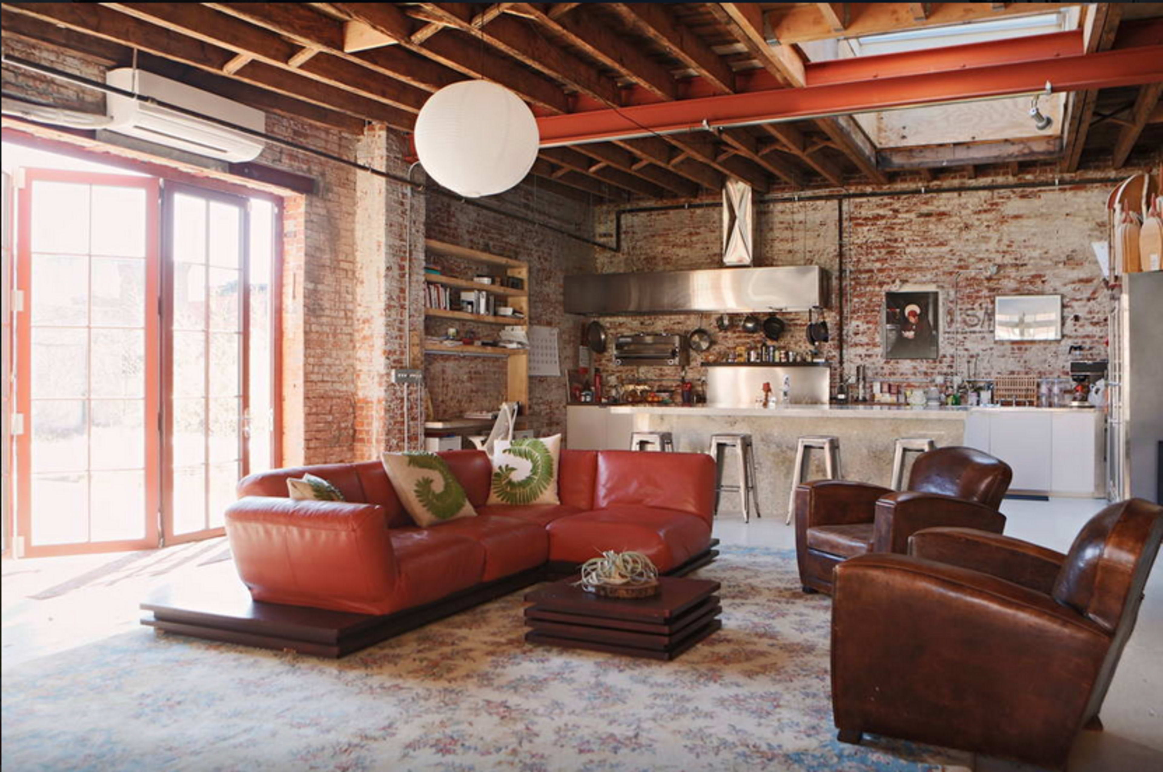 The image shows a spacious living area with exposed brick walls, a modern kitchen in the background, and leather furniture arranged around a large area rug.