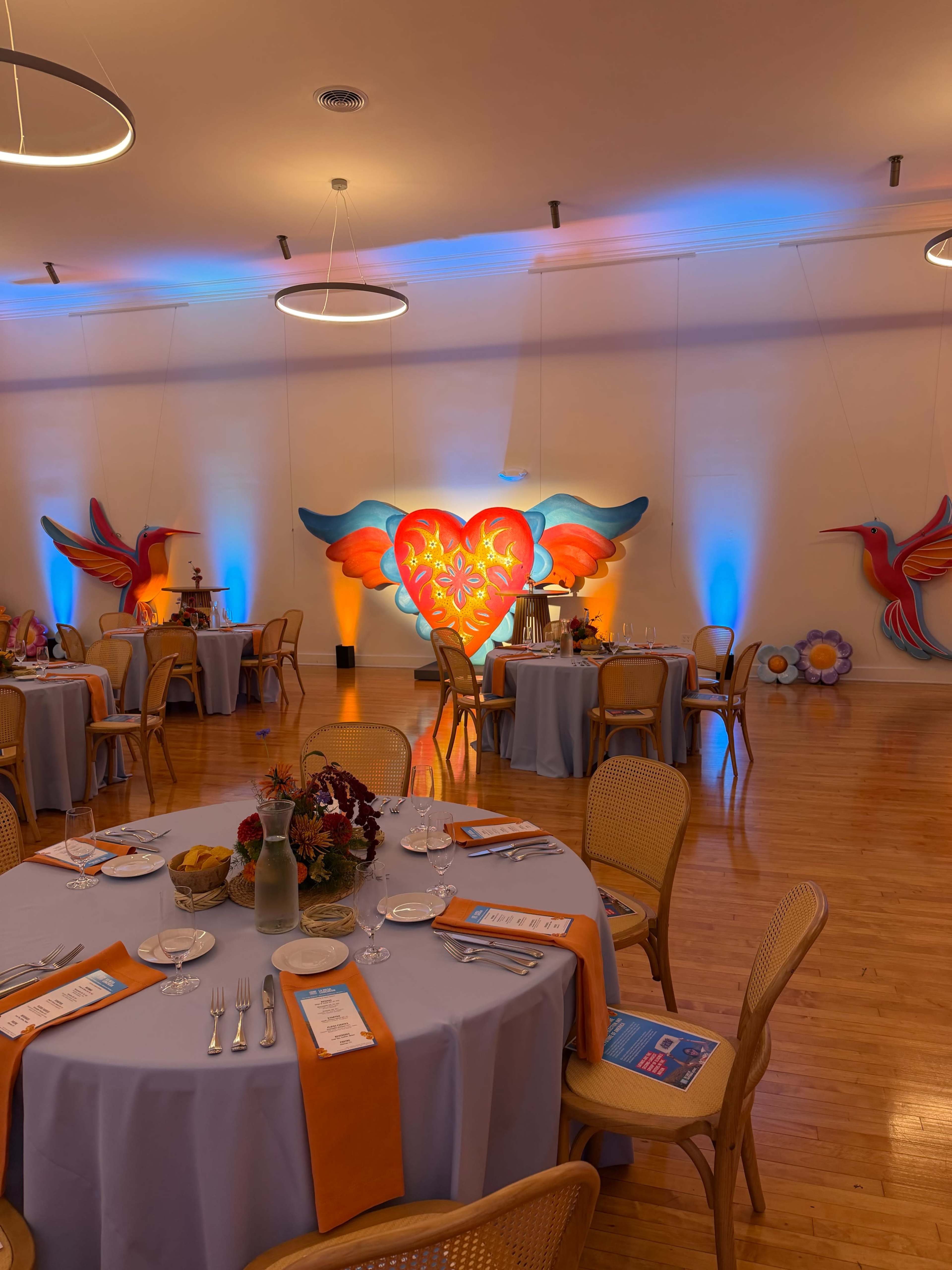 The image shows a banquet hall with round tables set for an event, featuring colorful decorations on the wall and soft blue and orange lighting.