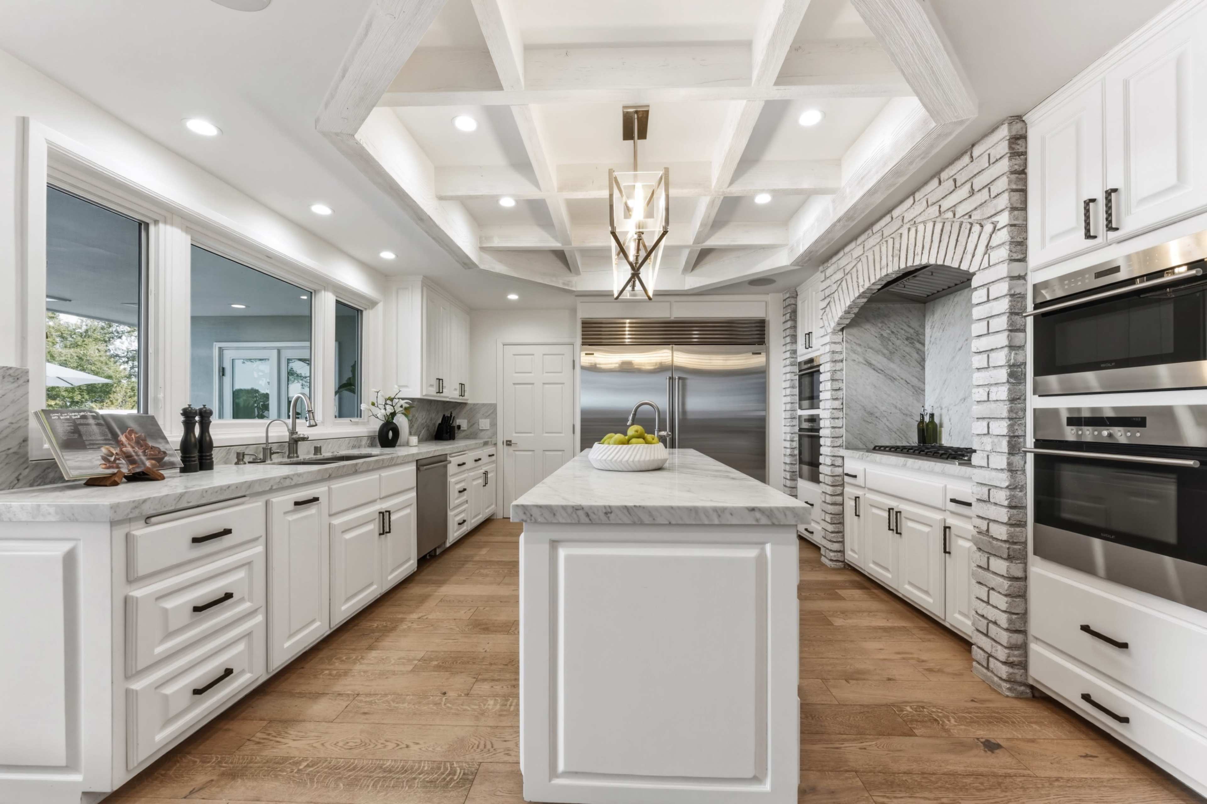 The image shows a modern kitchen featuring white cabinets, a large island with a marble countertop, stainless steel appliances, and wooden flooring.