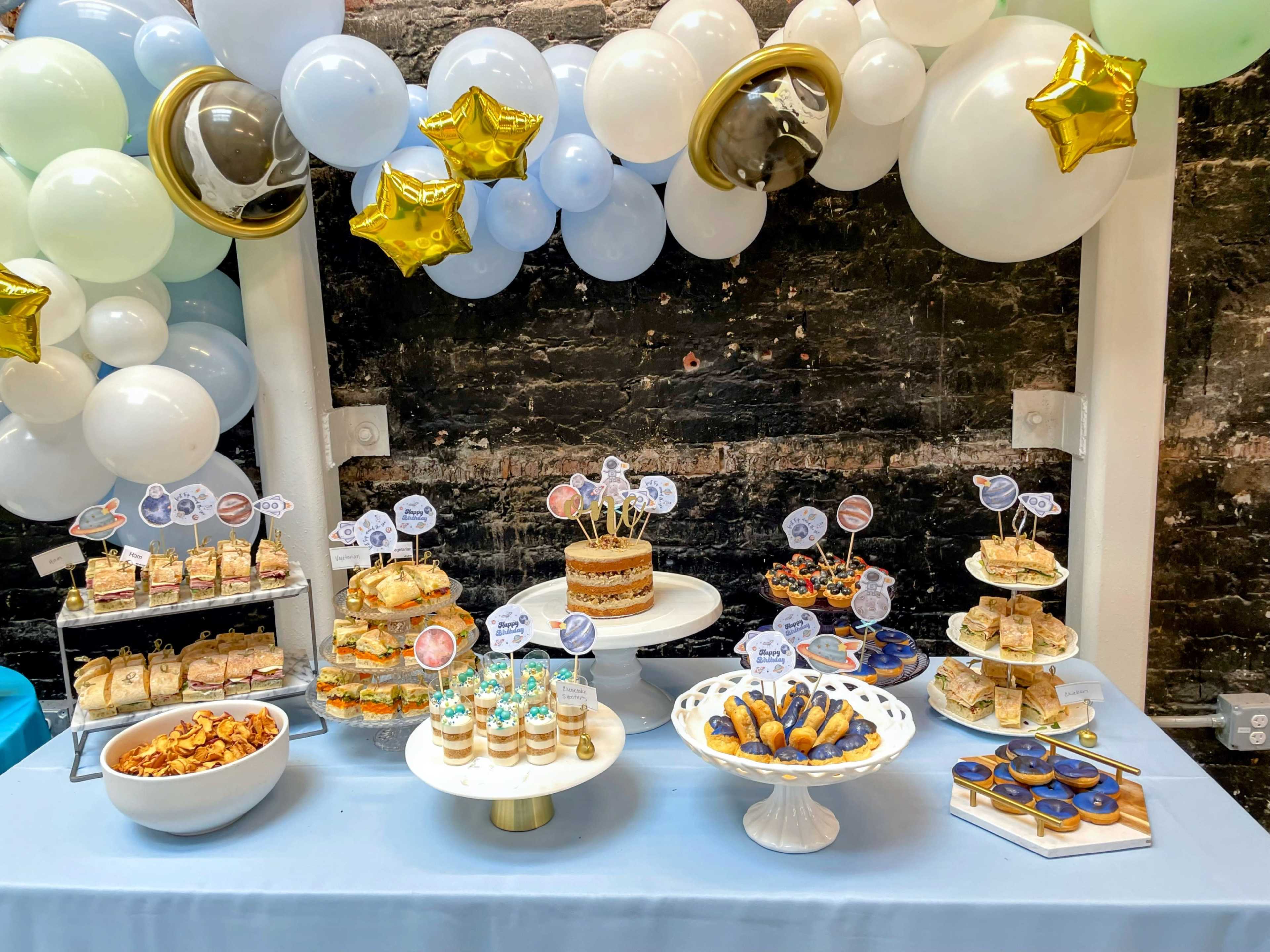 A dessert table is set up with a variety of treats, including a layered cake, pastries, and colorful desserts, surrounded by decorative balloons and star accents.