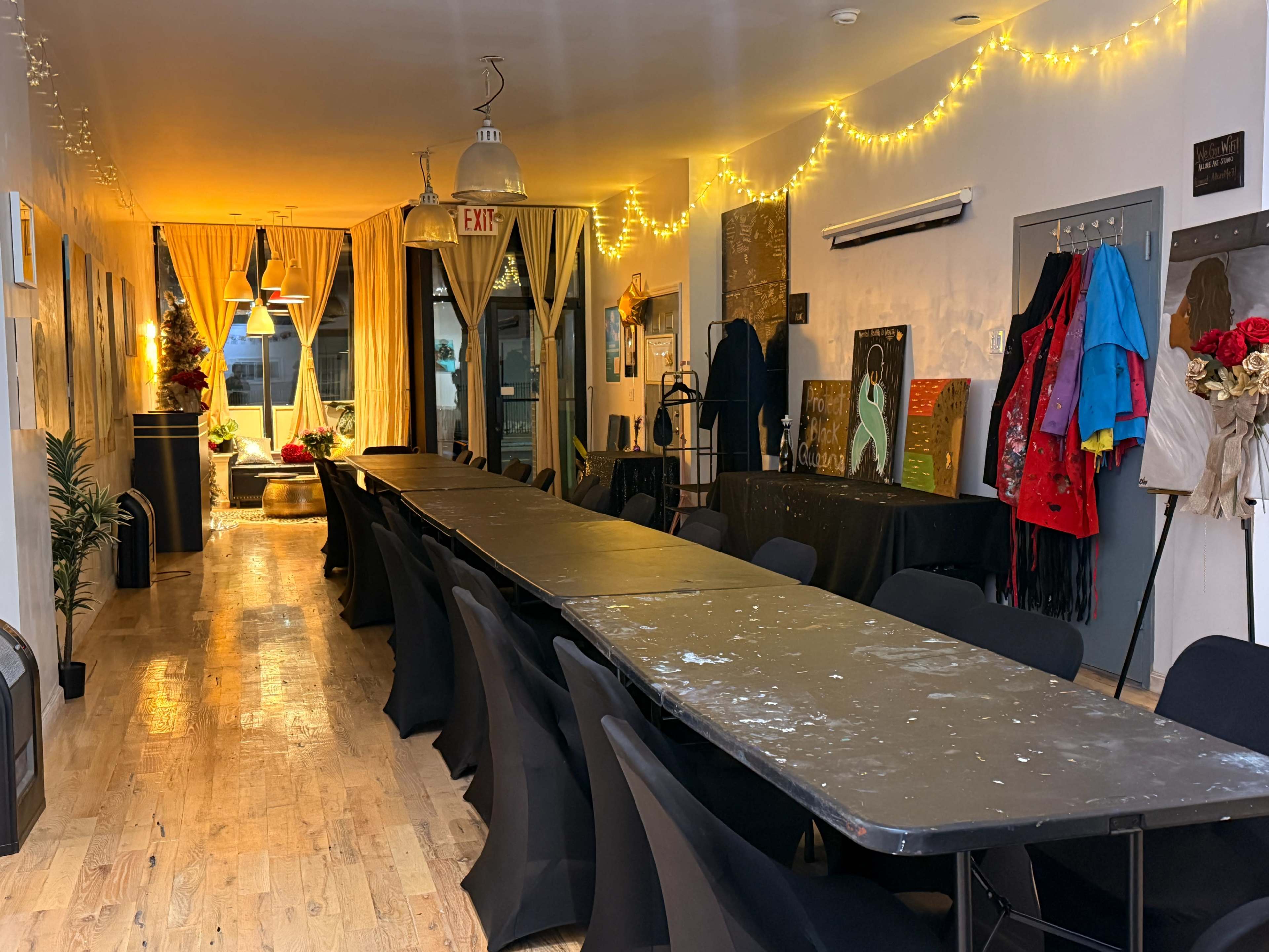 A long table is set up in a dimly lit room adorned with string lights, with chairs arranged neatly around it and artwork displayed on the walls.