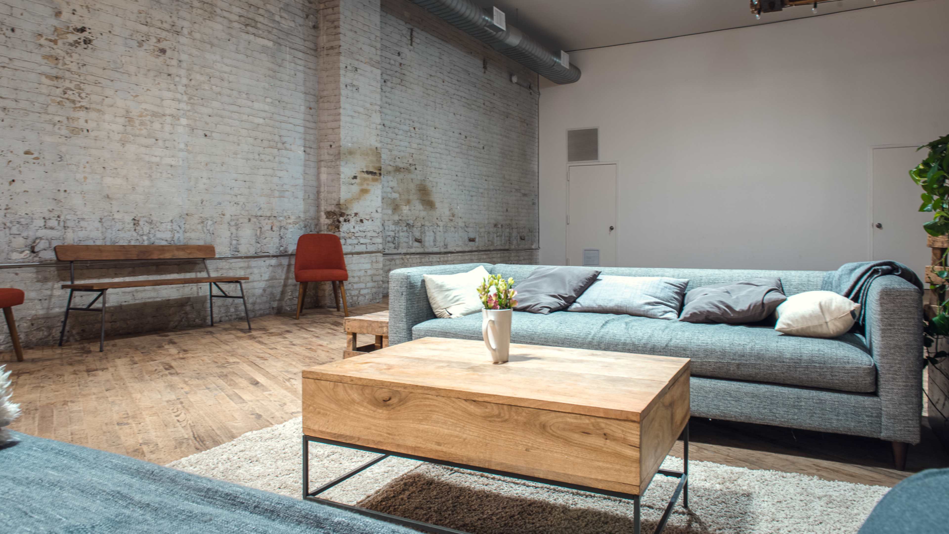 A spacious, minimalistic living area with a gray sofa, a wooden coffee table, and a small table with chairs against a white brick wall.