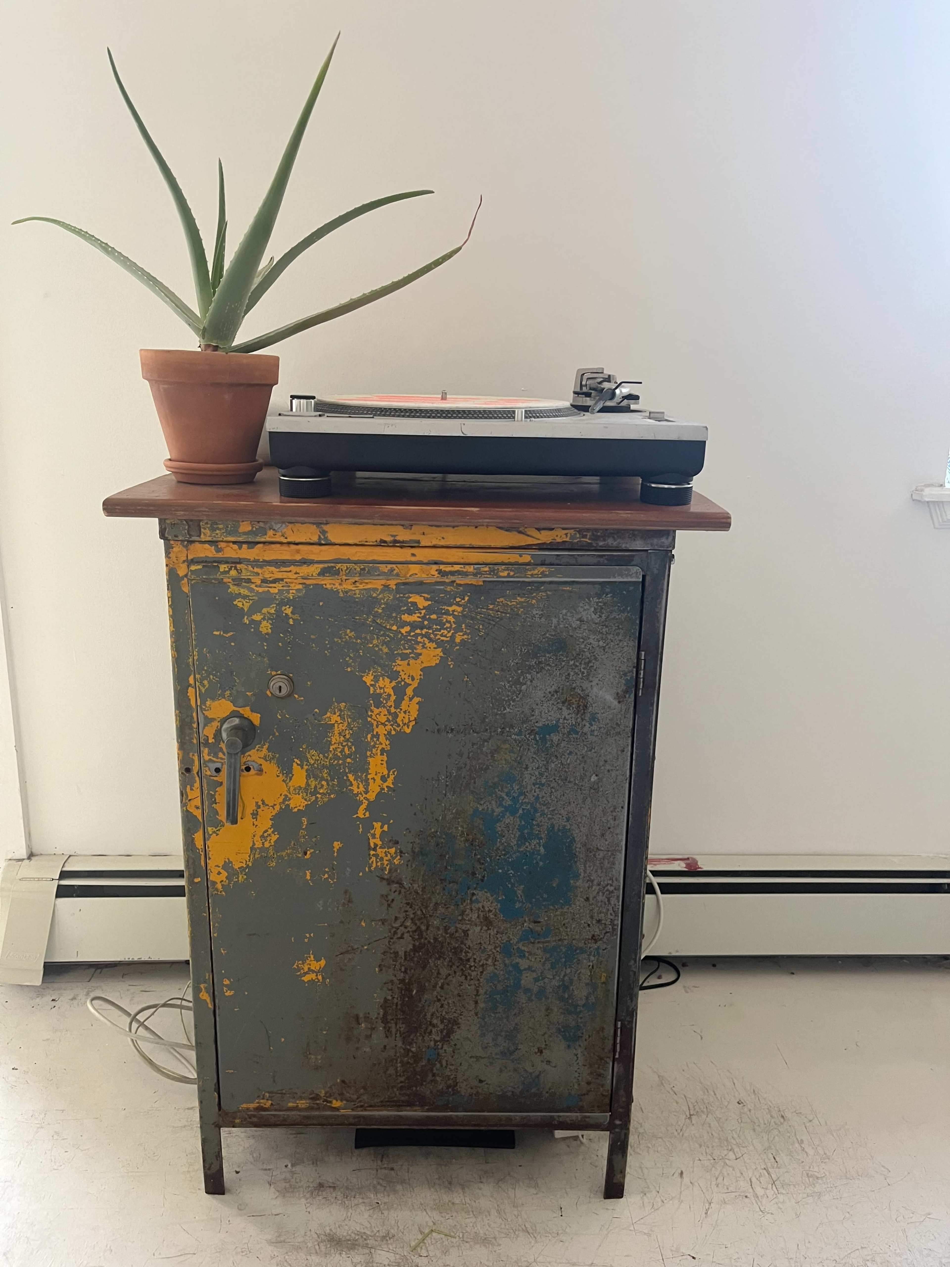 A vintage cabinet with a weathered finish holds a turntable on its top, while a potted aloe vera plant sits beside it.