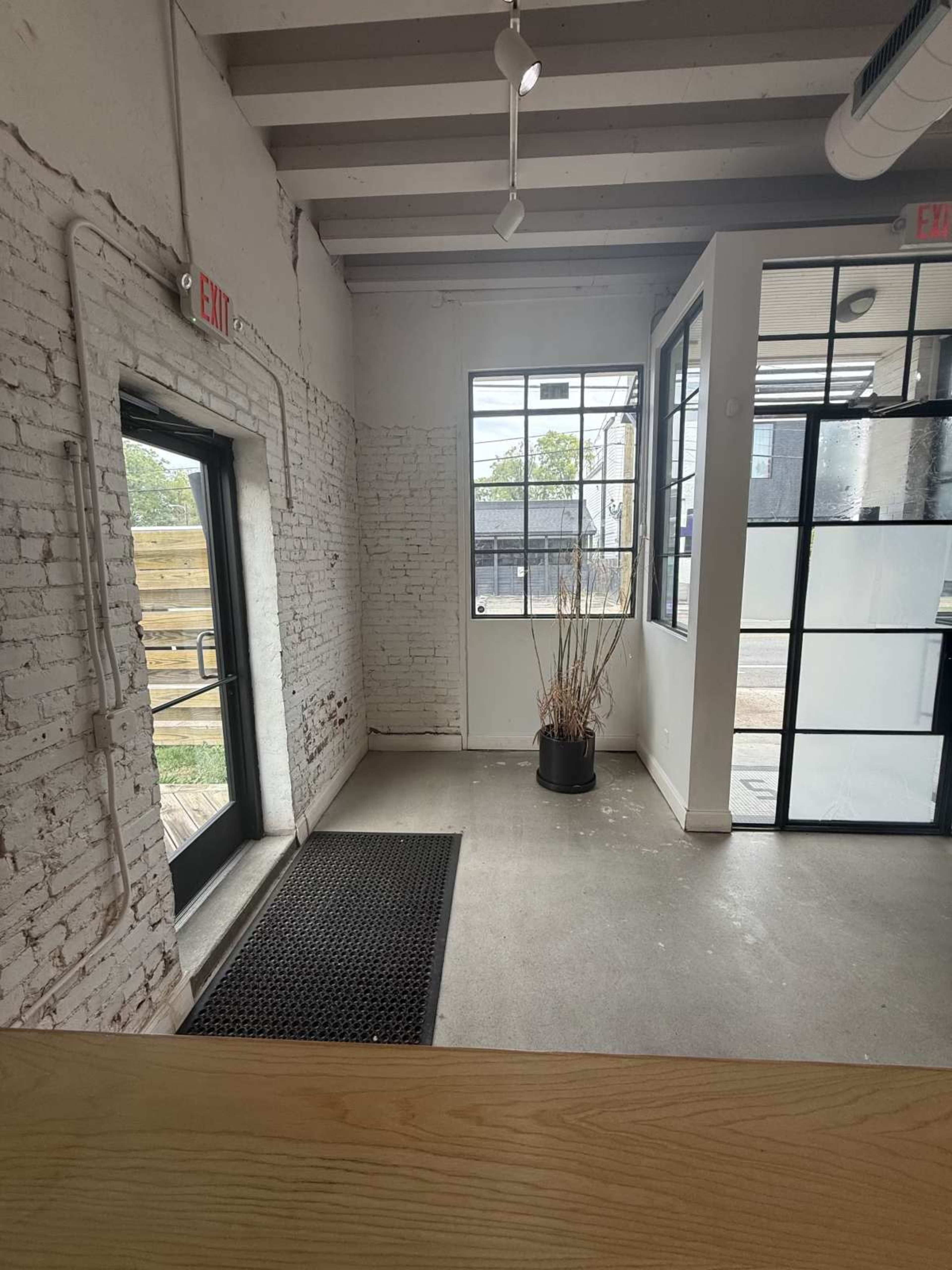 The image shows a sparse interior space with white brick walls, a large window, a door leading outside, and a planter with bare branches.