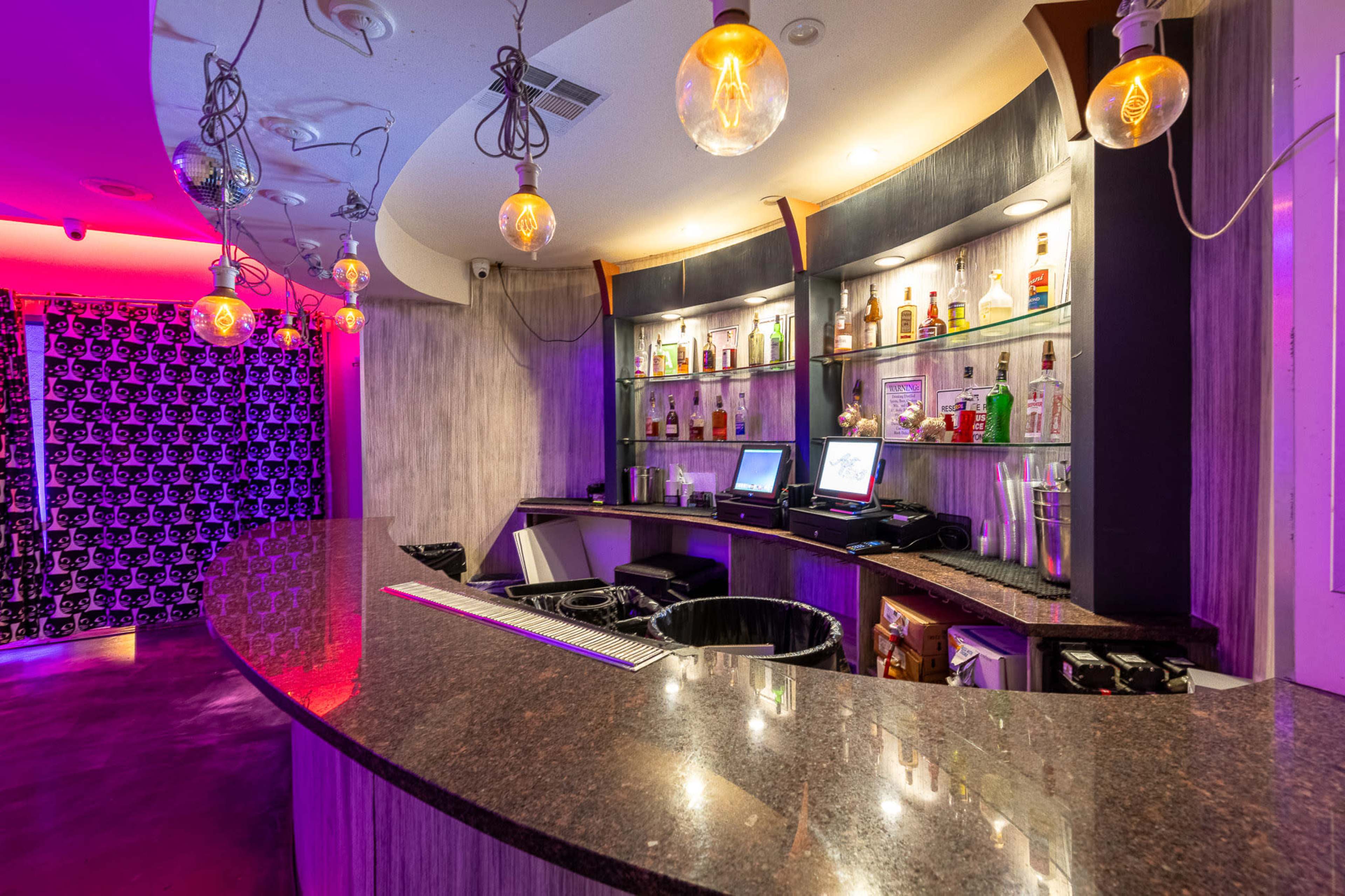 The image shows a modern bar with a curved countertop, illuminated shelves displaying various bottles, and pendant lighting in a dimly lit environment.