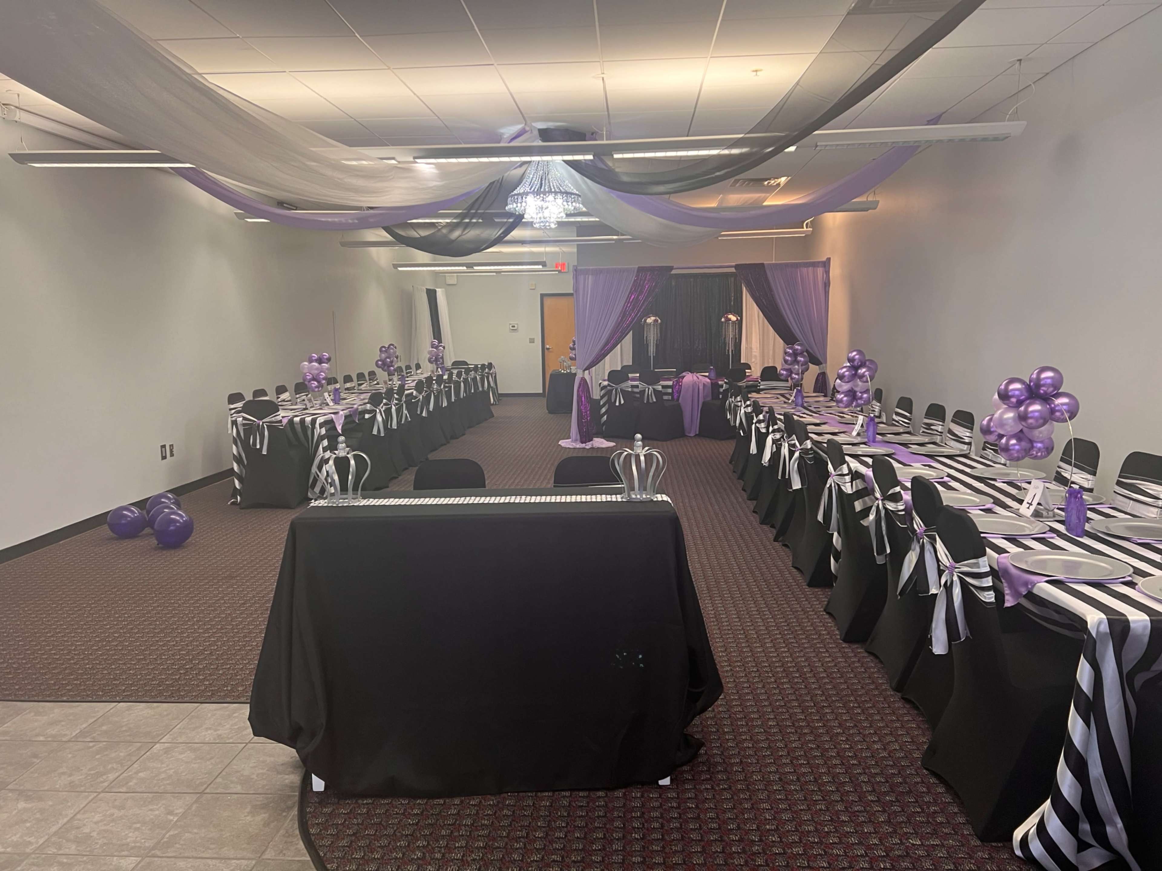 A decorated event space with black and purple table settings, balloons, and draped fabric overhead.
