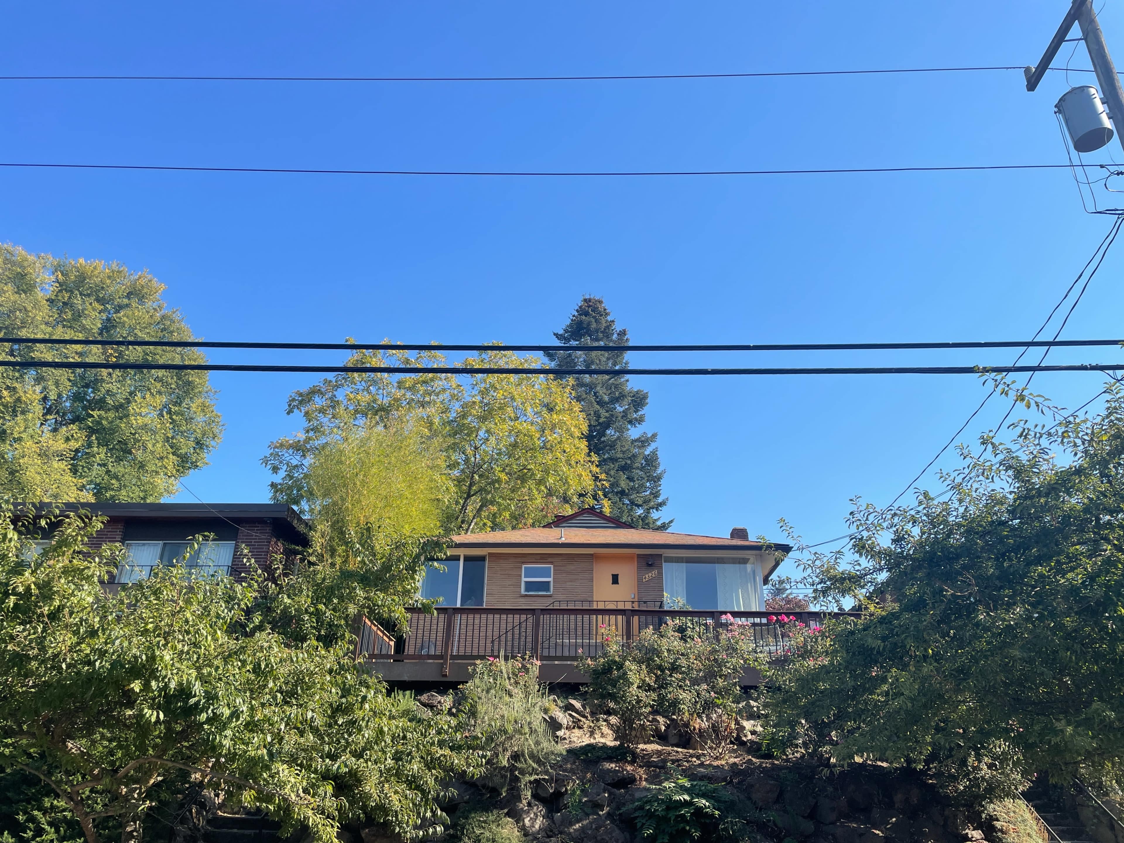A house stands on a hillside, surrounded by trees and power lines against a clear blue sky.