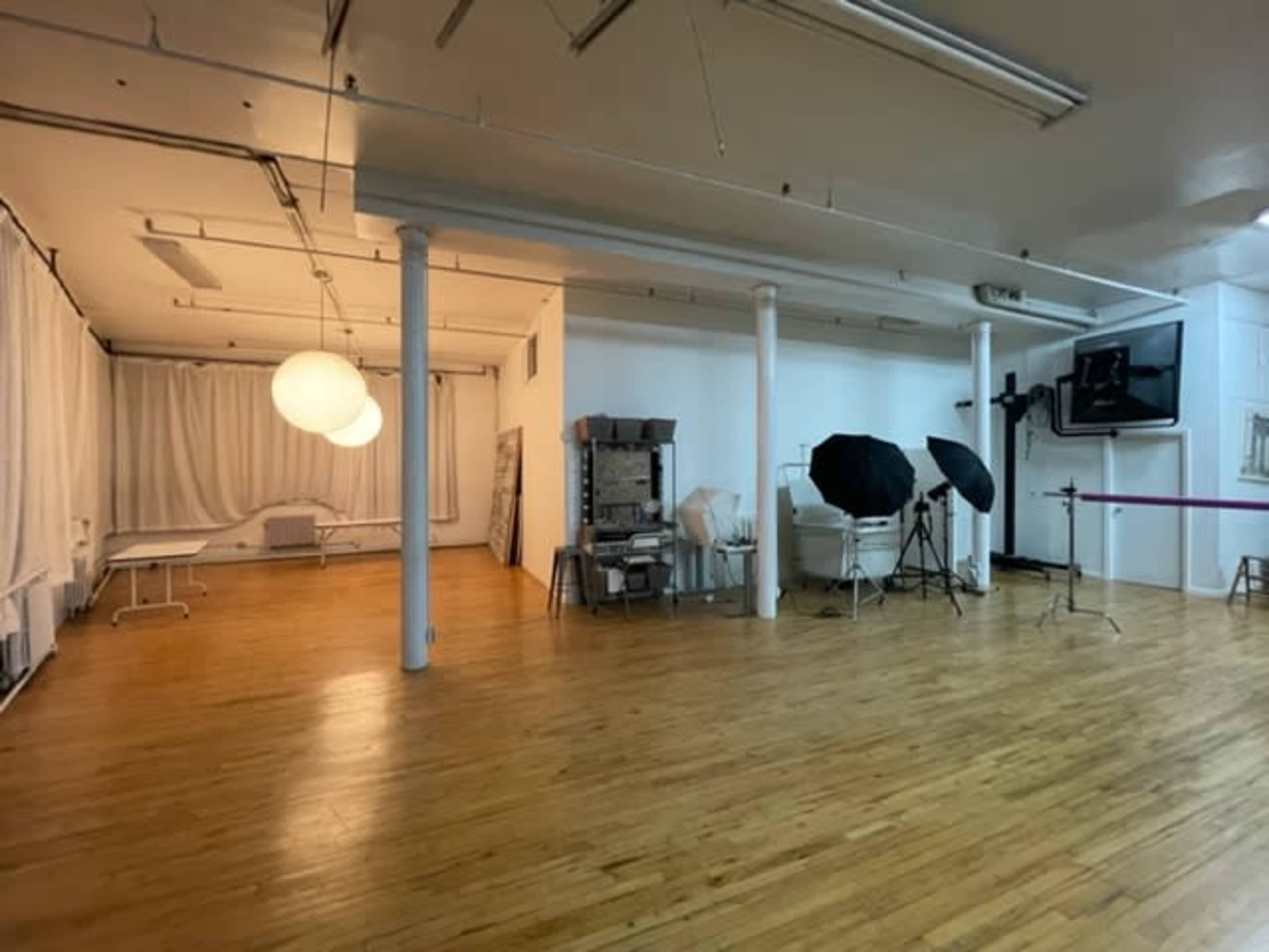 The image shows a spacious studio with wooden floors, white drapes, lighting equipment, and a few chairs arranged for a photography setup.