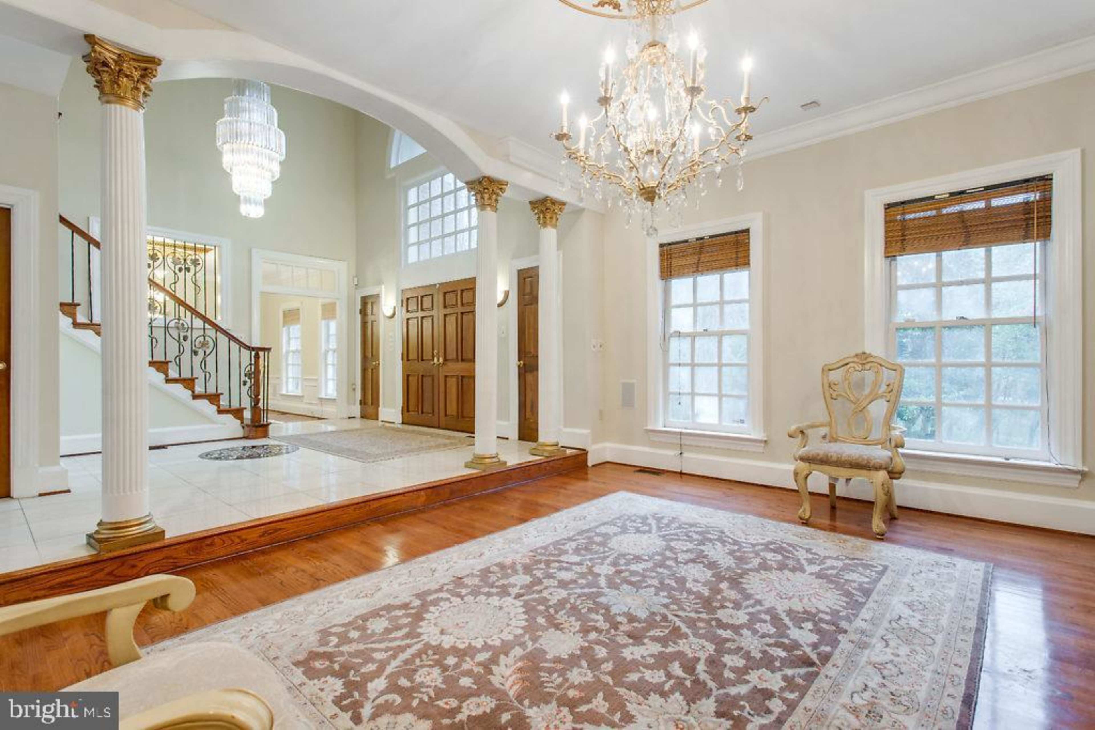 A spacious foyer with a chandelier, wooden floors, and a decorative rug, leading to a staircase and double front doors.