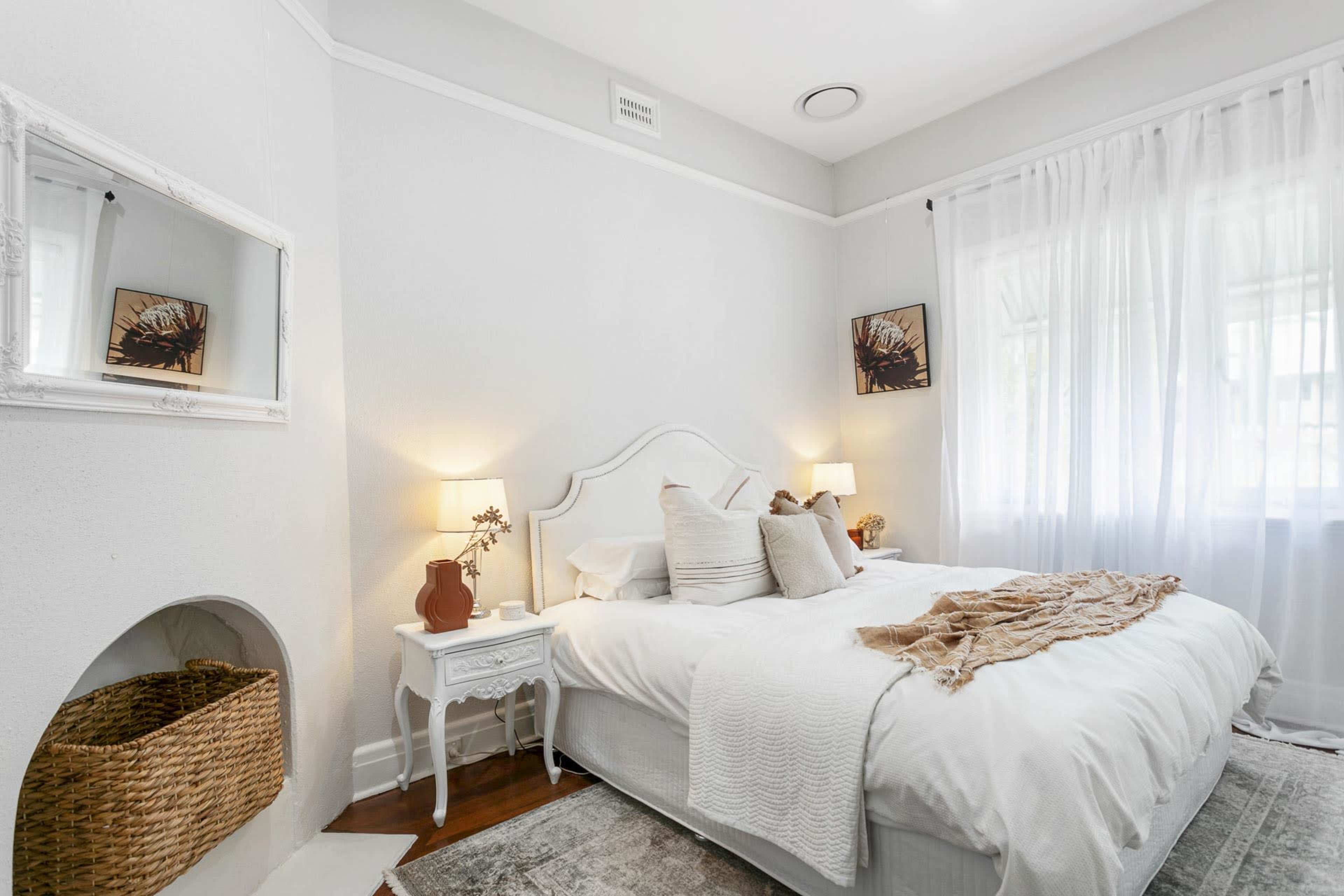 A bright bedroom features a white bed with decorative pillows, two bedside lamps, and a wicker basket beside a small table, all set against light-colored walls and large windows.