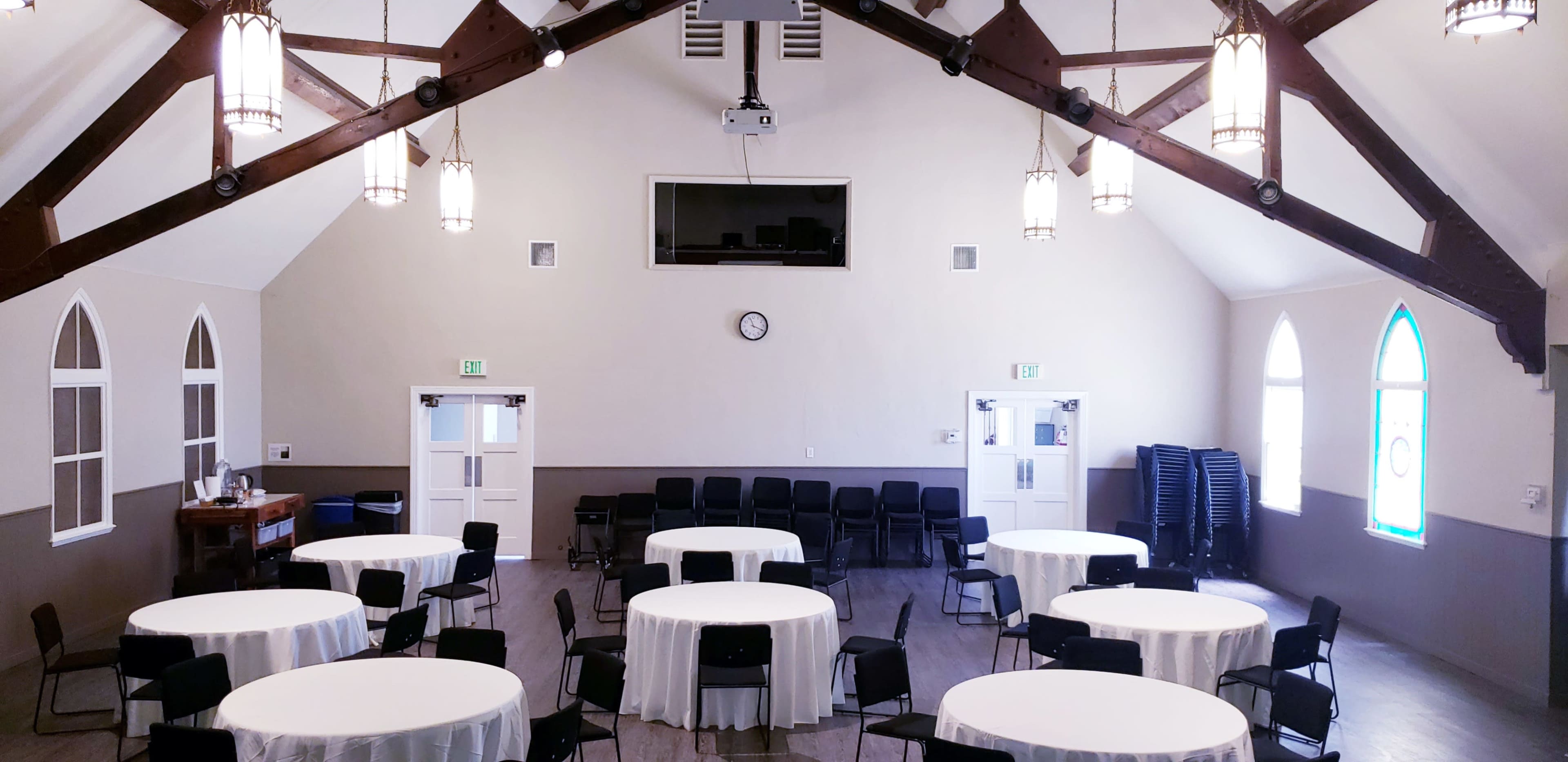 The interior of a spacious hall features tables with white tablecloths, black chairs arranged in rows, and a projector mounted on the ceiling.