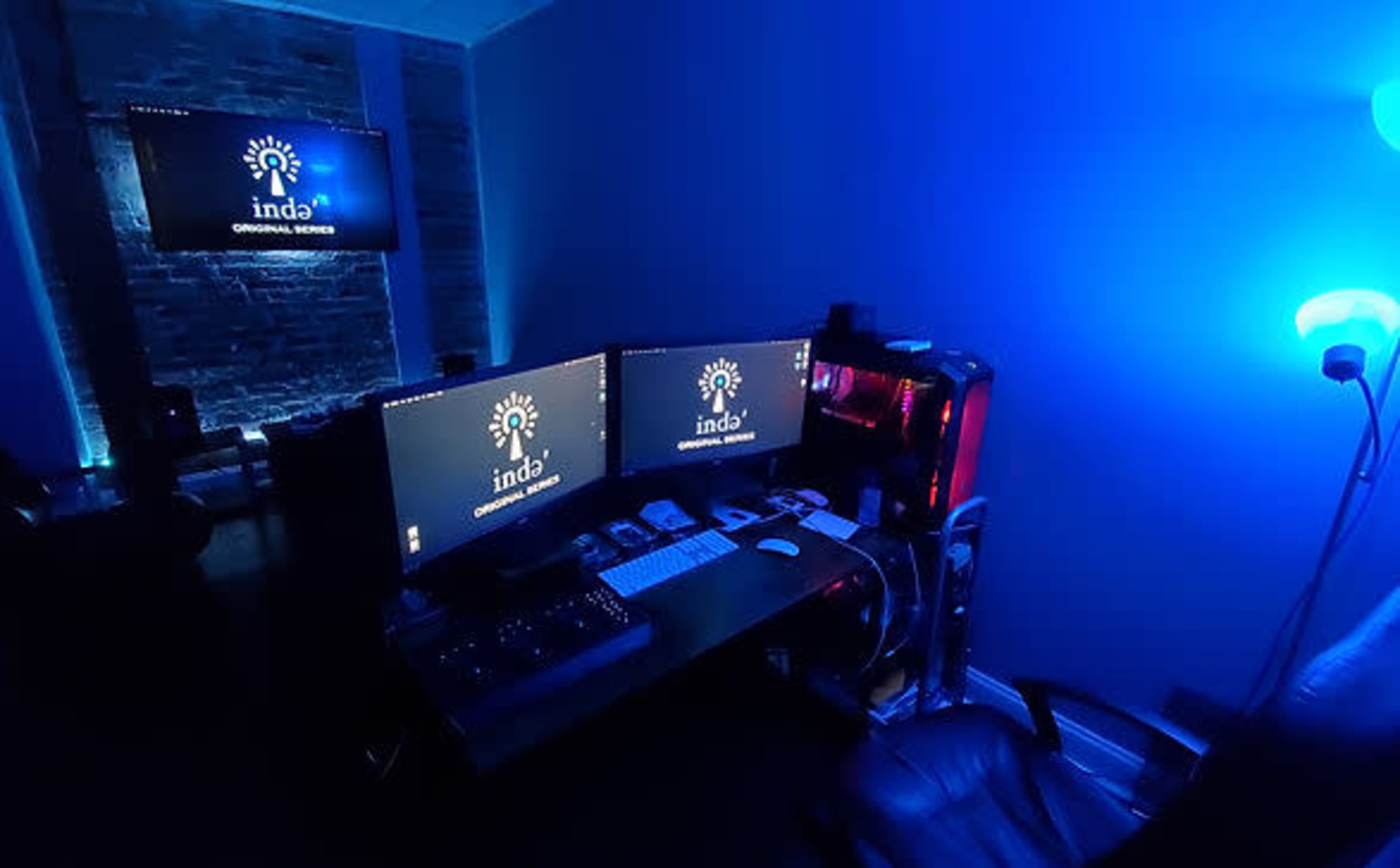 A dimly lit workspace featuring two computer monitors, a modern gaming PC, and ambient blue lighting.