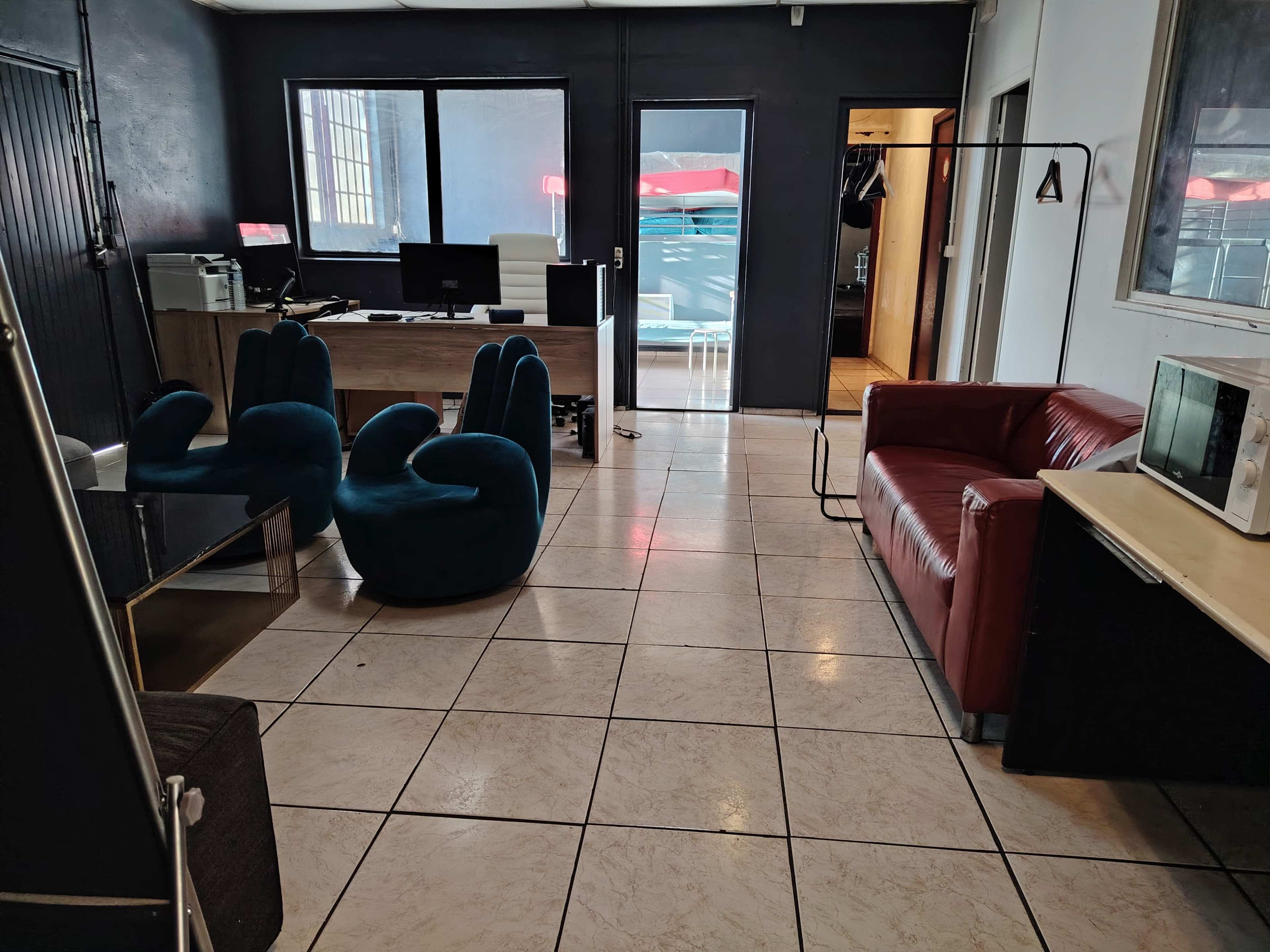 The image shows a modern office space with a mix of dark and light furniture, featuring a red sofa, blue chairs, a desk with a computer, and a glass door leading to an outdoor area.