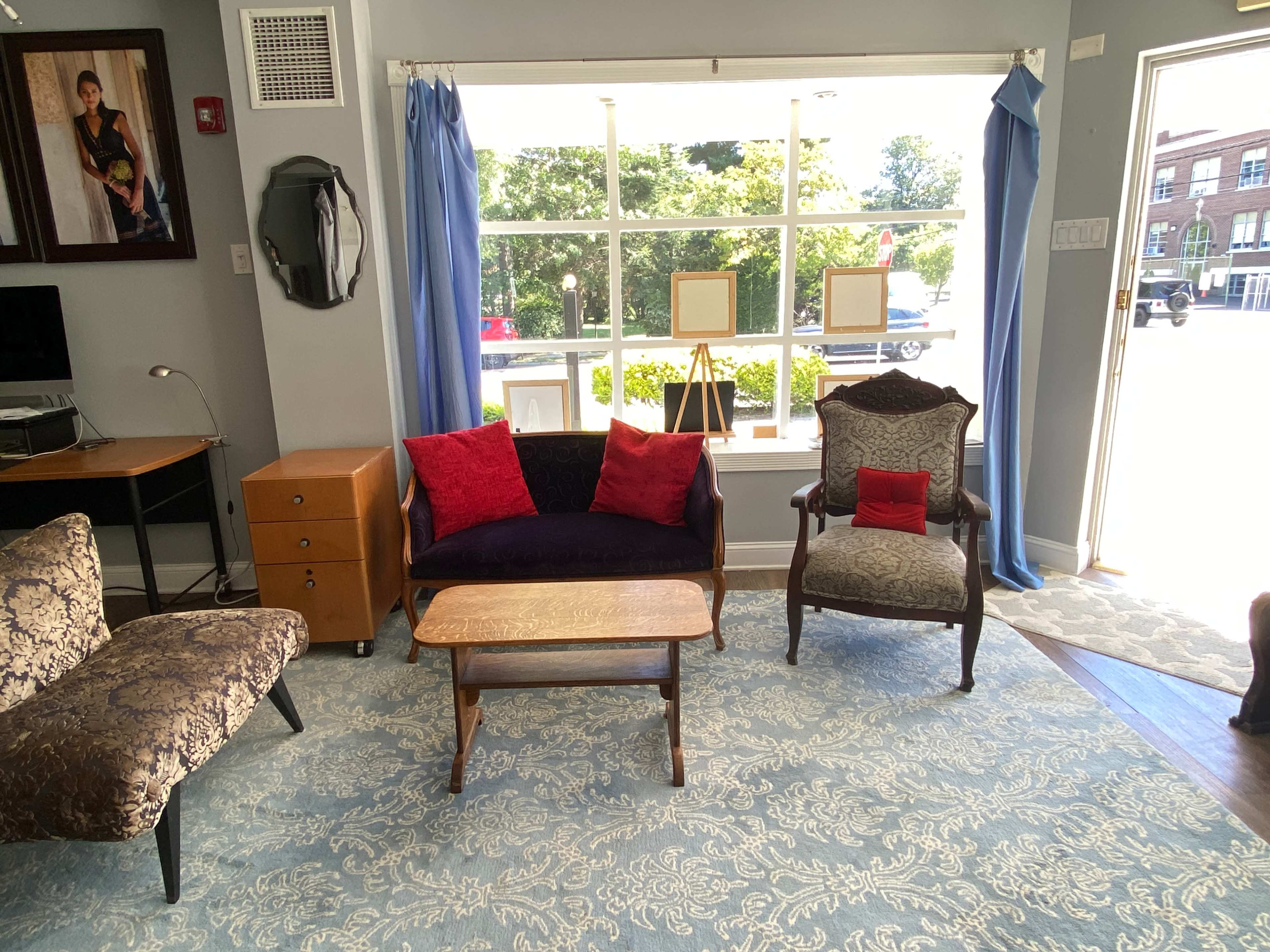 The image shows a small seating area with a patterned rug, a wooden coffee table, and three upholstered chairs, situated near a large window.