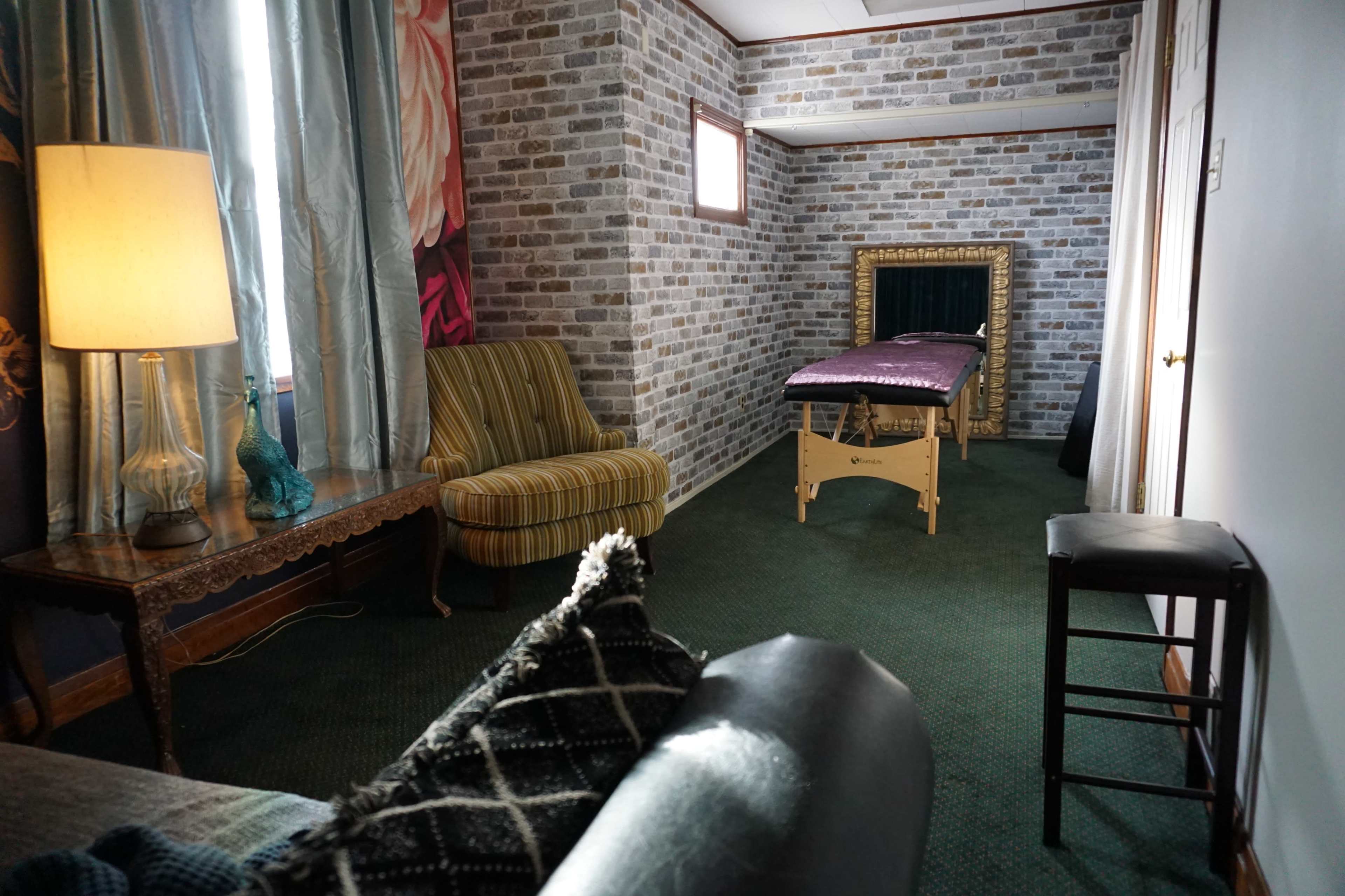 A room with a massage table against a brick-patterned wall, a striped armchair, a wooden table, and a stool, all illuminated by a standing lamp.