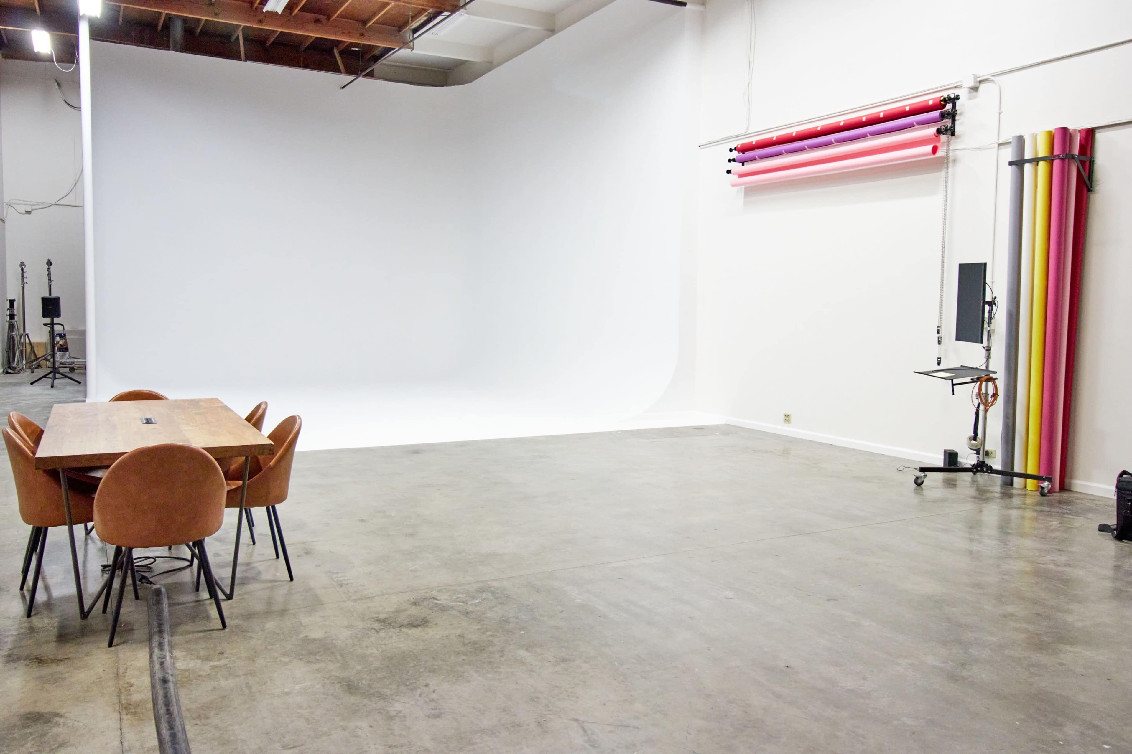 A spacious photography studio with a white backdrop, a wooden table surrounded by chairs, and colorful backdrop rolls on the wall.
