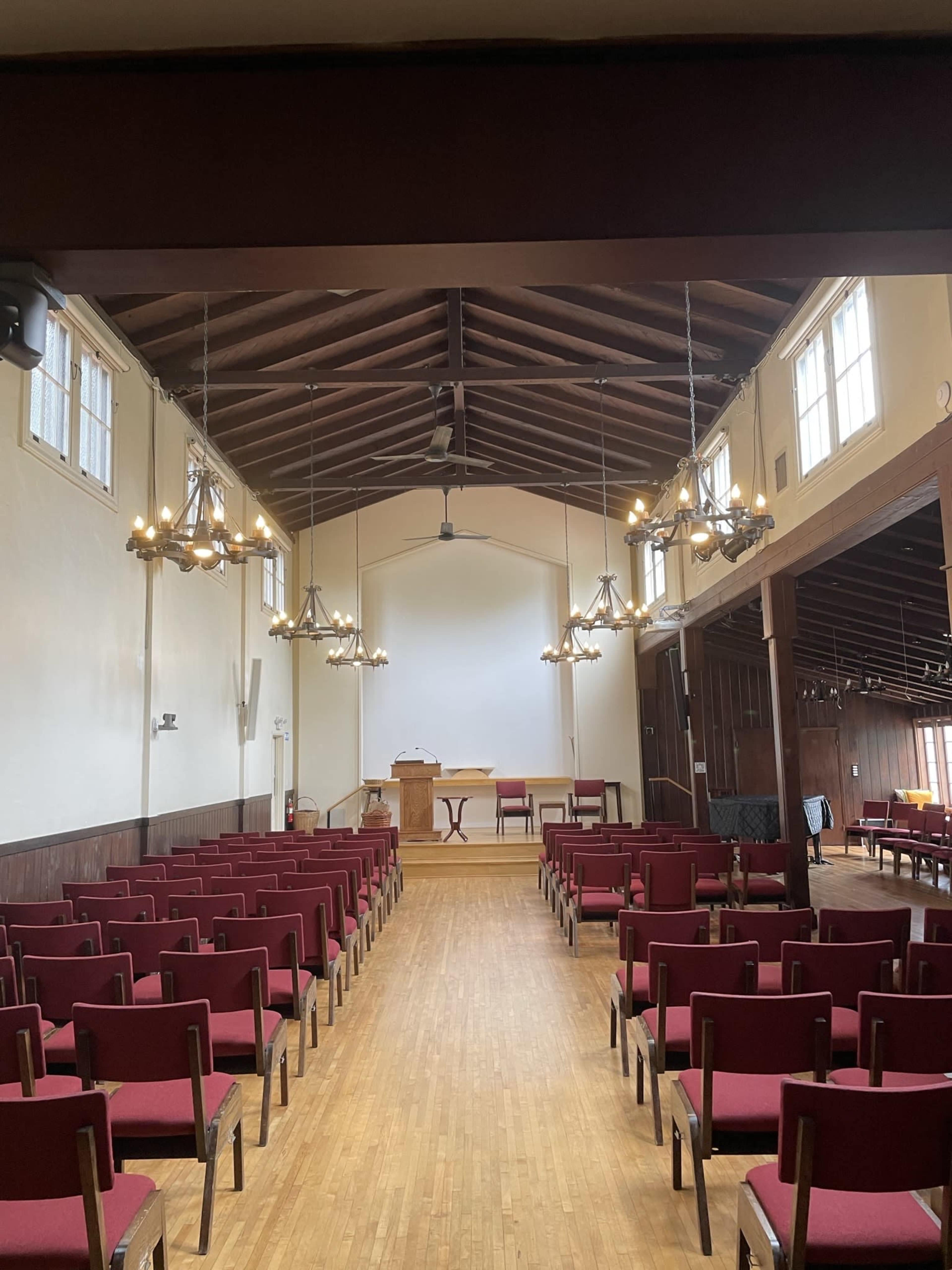 A spacious chapel with rows of red chairs facing a wooden podium and a table under a bright, high ceiling.