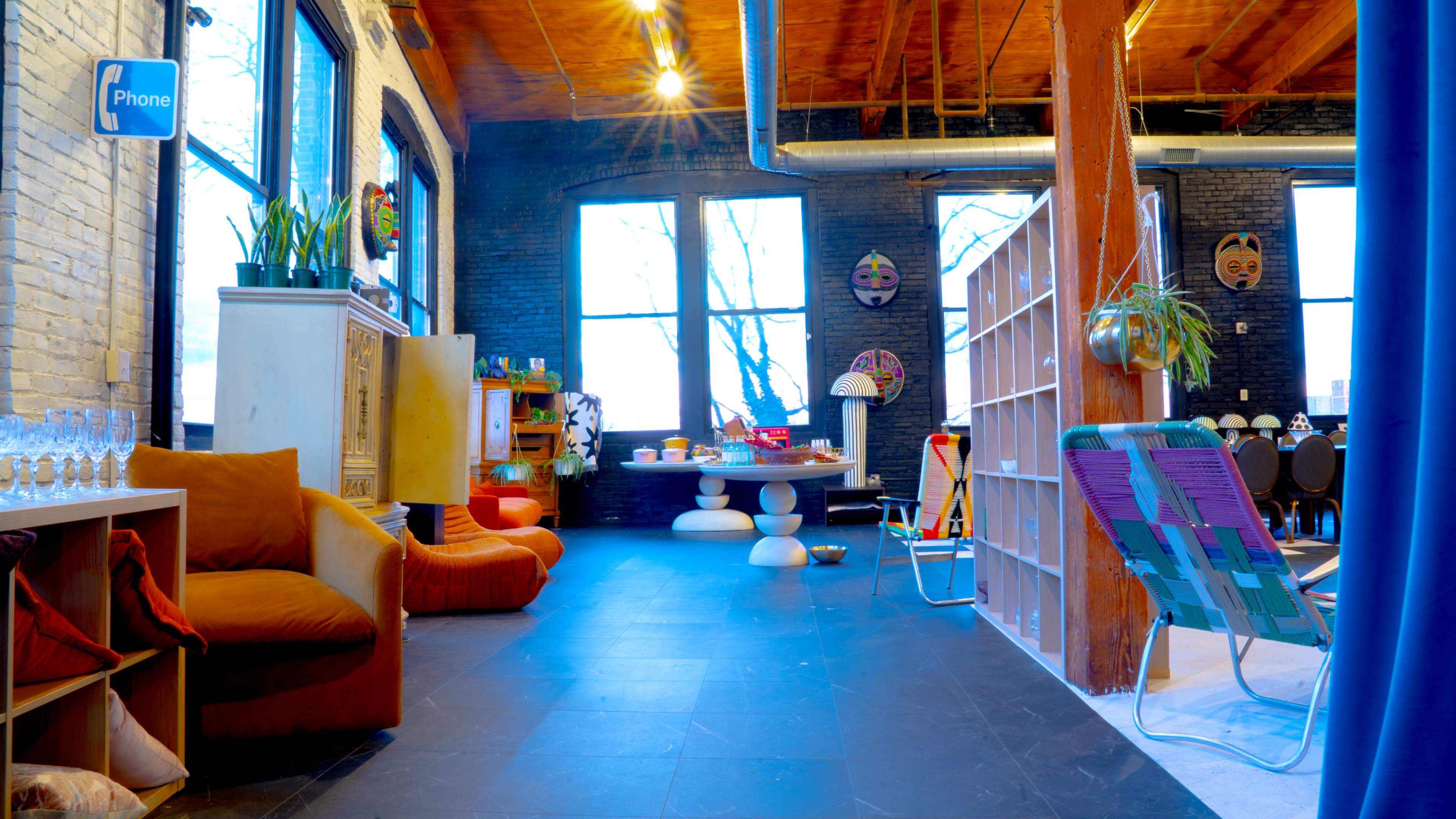 The image shows a spacious interior room with large windows, featuring colorful furniture, a dining table, and decorative masks on the walls.