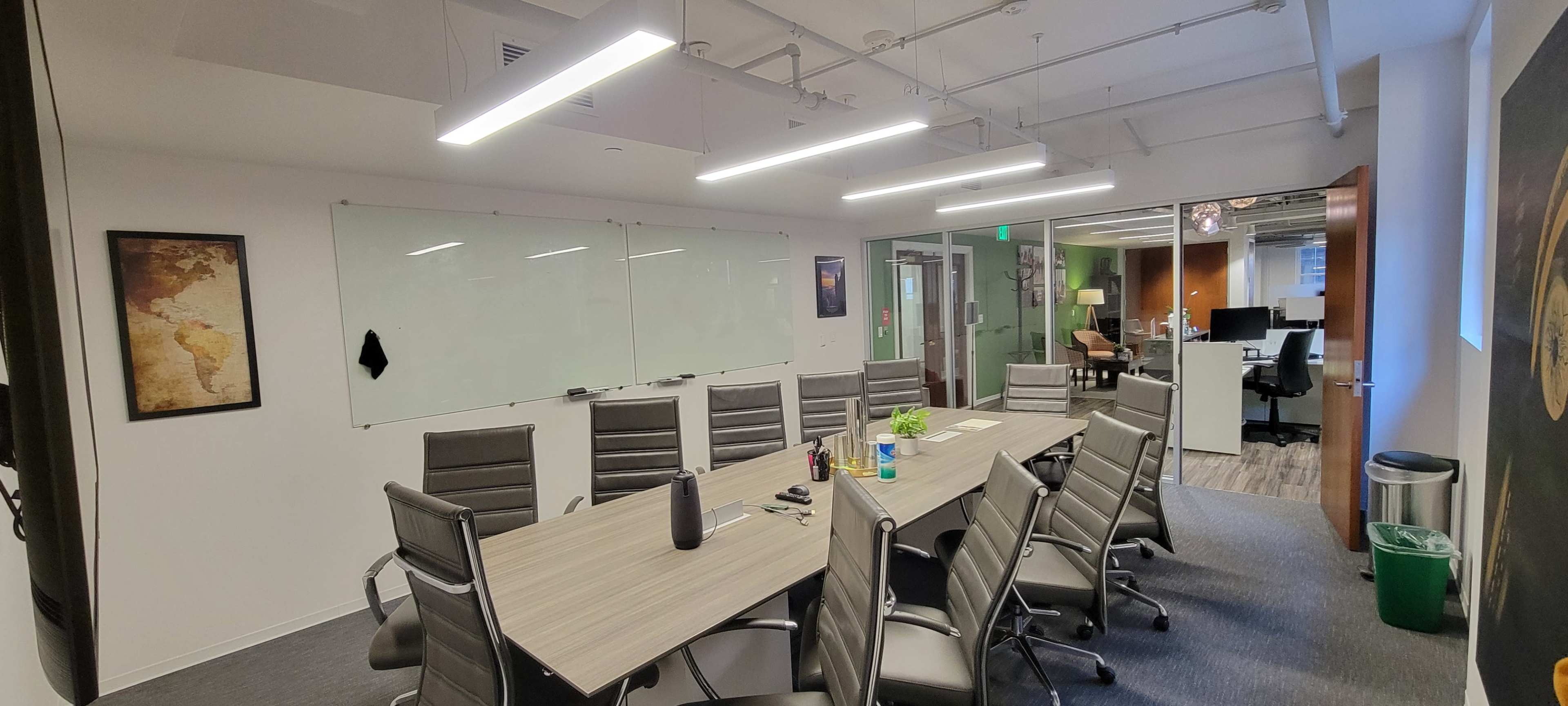 A conference room features a long wooden table surrounded by ergonomic chairs, with a whiteboard and glass wall in the background.