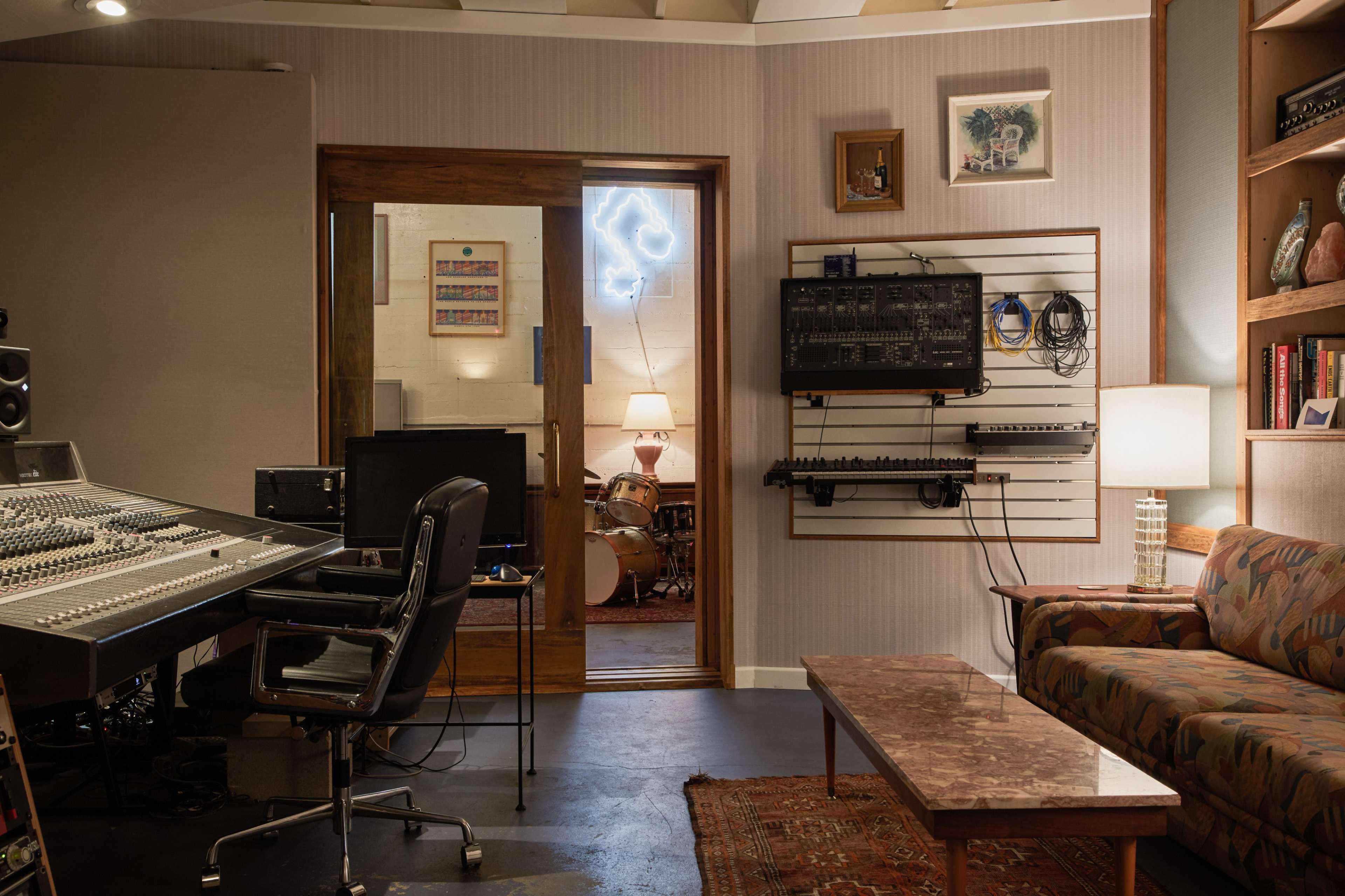 The image shows a music recording studio featuring a mixing console, a computer desk, and a view into a room with a drum set and a cozy seating area.