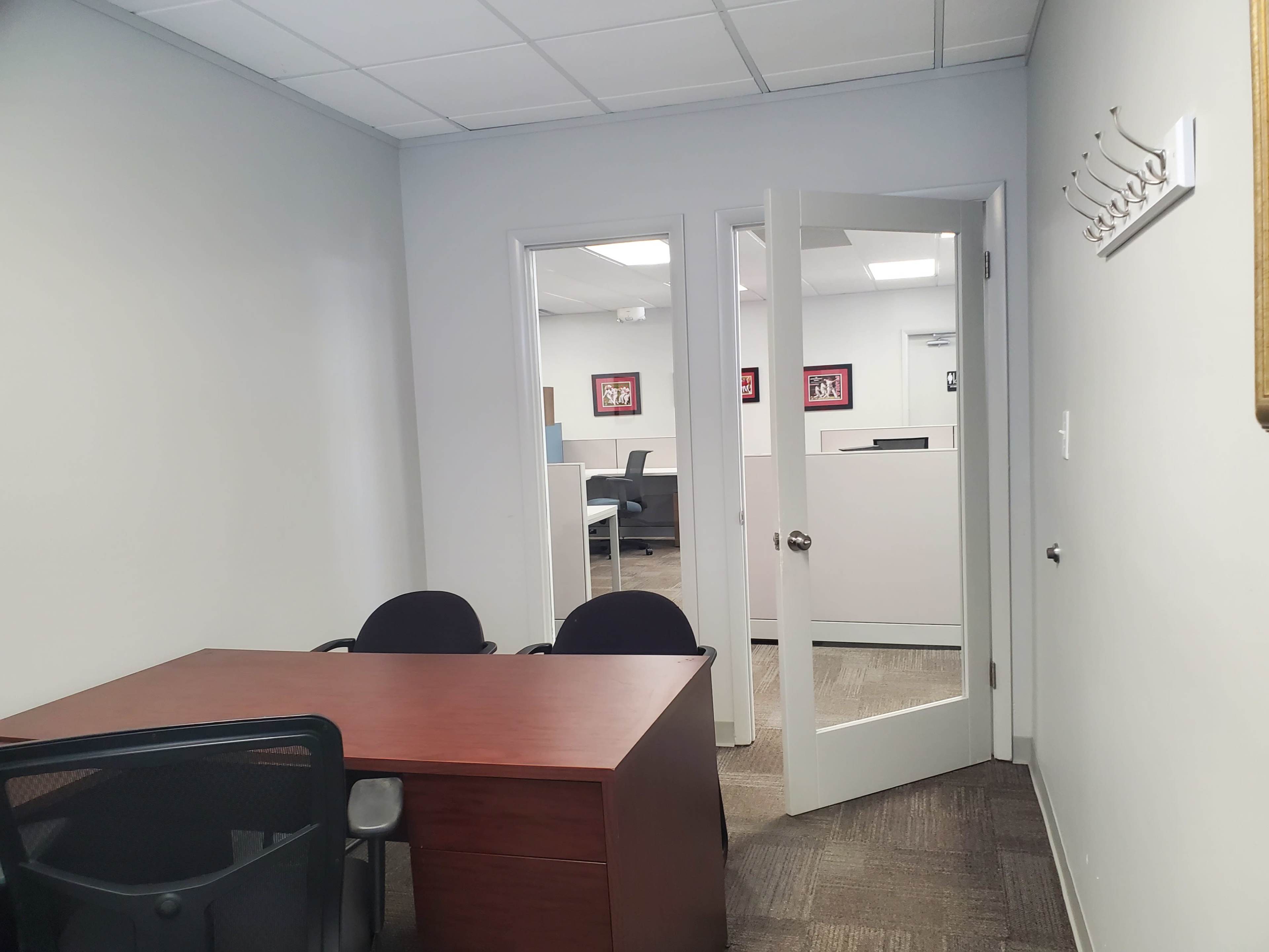 A small office space with a wooden desk, two black chairs, and an open doorway leading to another area with cubicles.