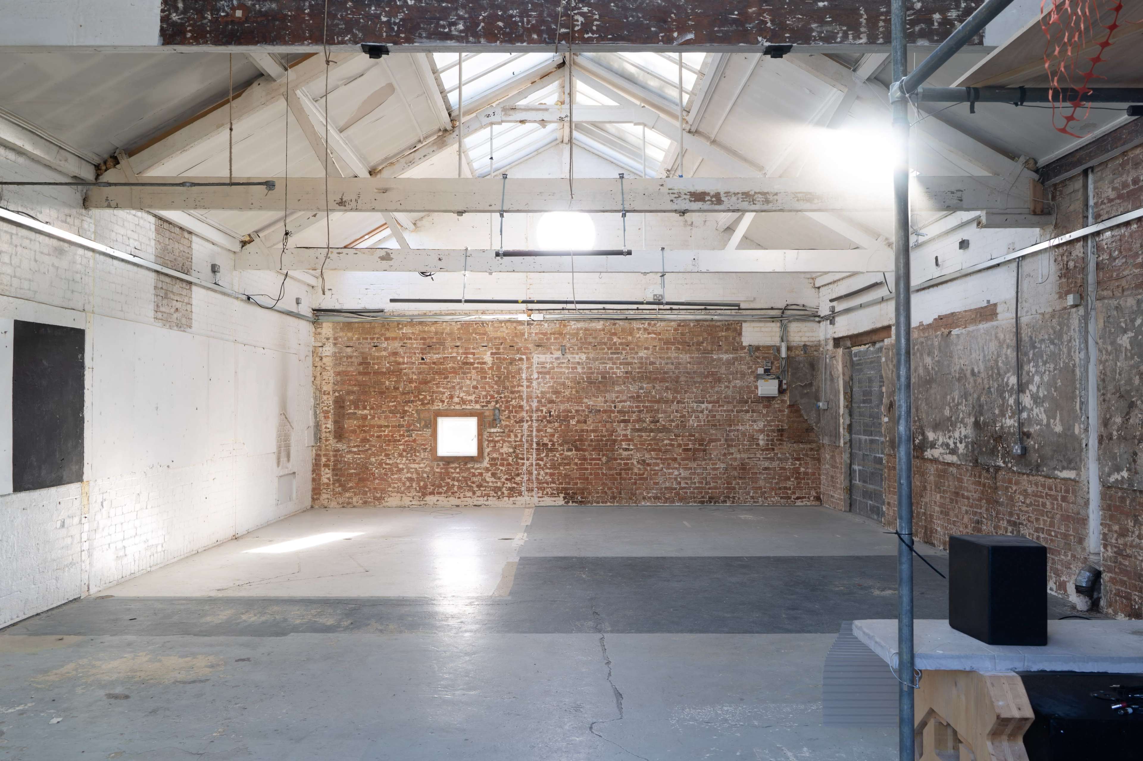The image shows a spacious, industrial-style room with exposed brick walls, large beams, and concrete flooring illuminated by natural light from skylights.