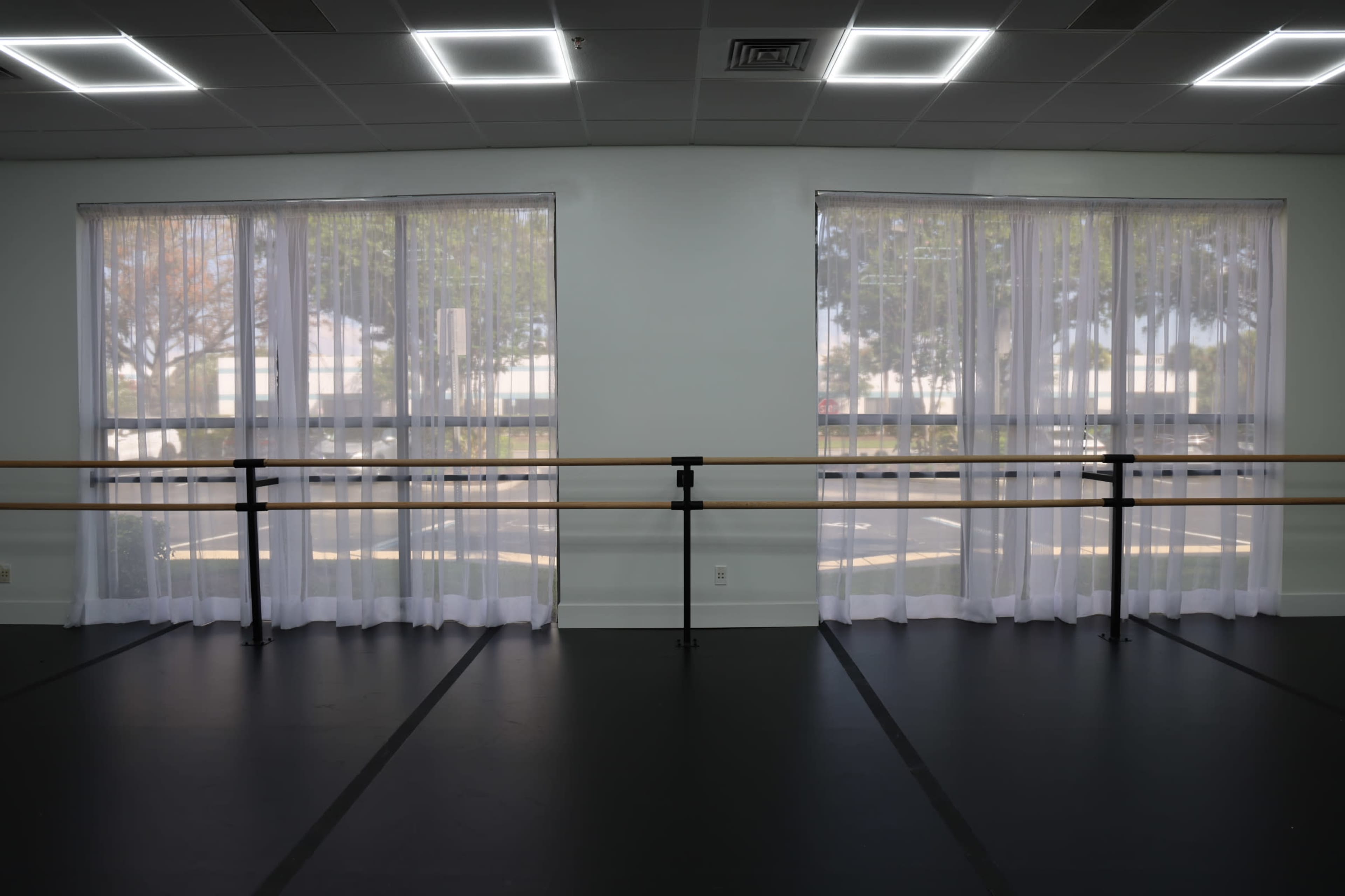 The image shows a dance studio with large windows dressed in sheer curtains and wooden ballet barres positioned against the walls.