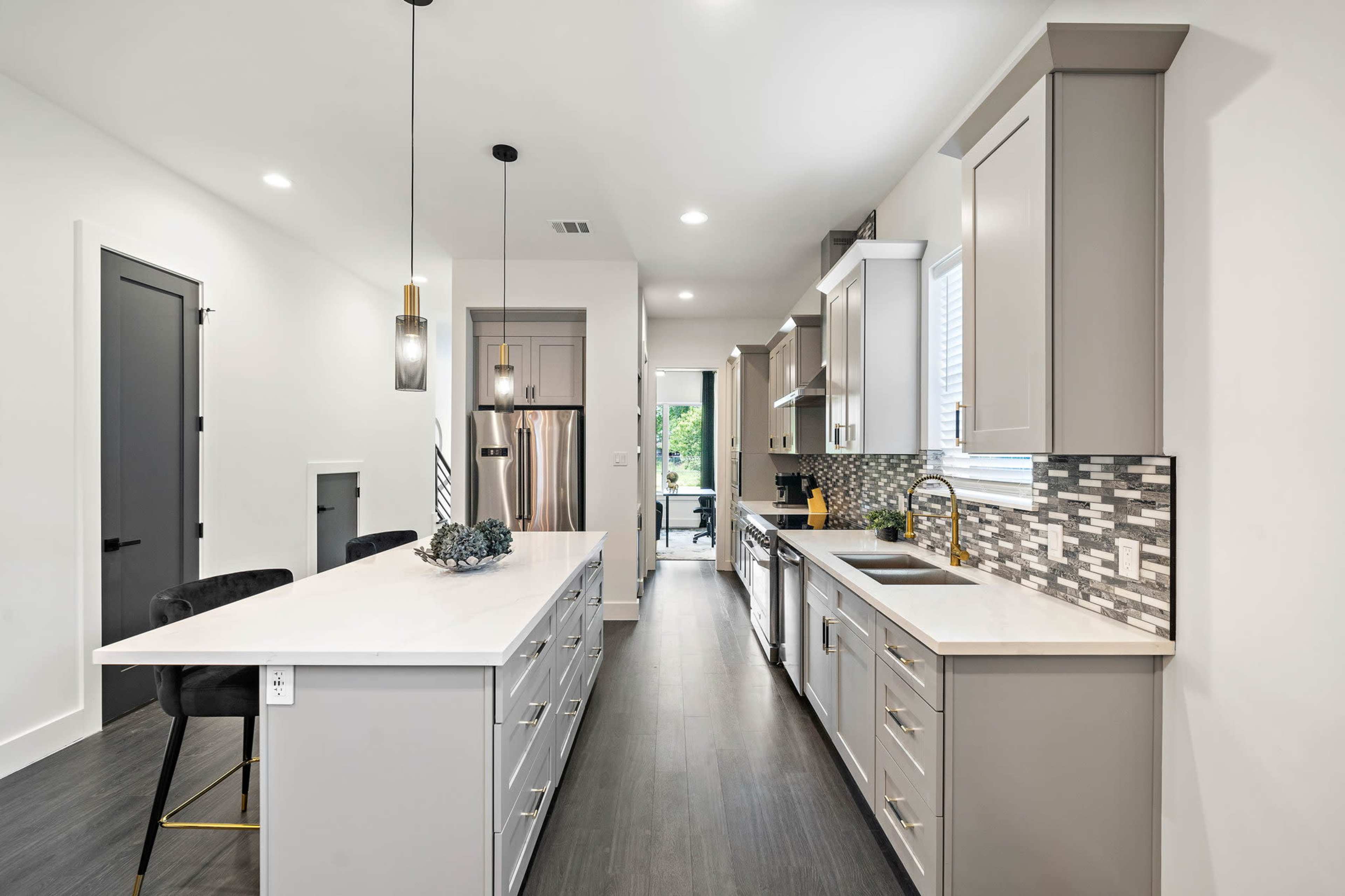A modern kitchen features a central island with a light-colored countertop, gray cabinetry, and a mosaic tile backsplash, leading to a doorway that opens to an outdoor view.