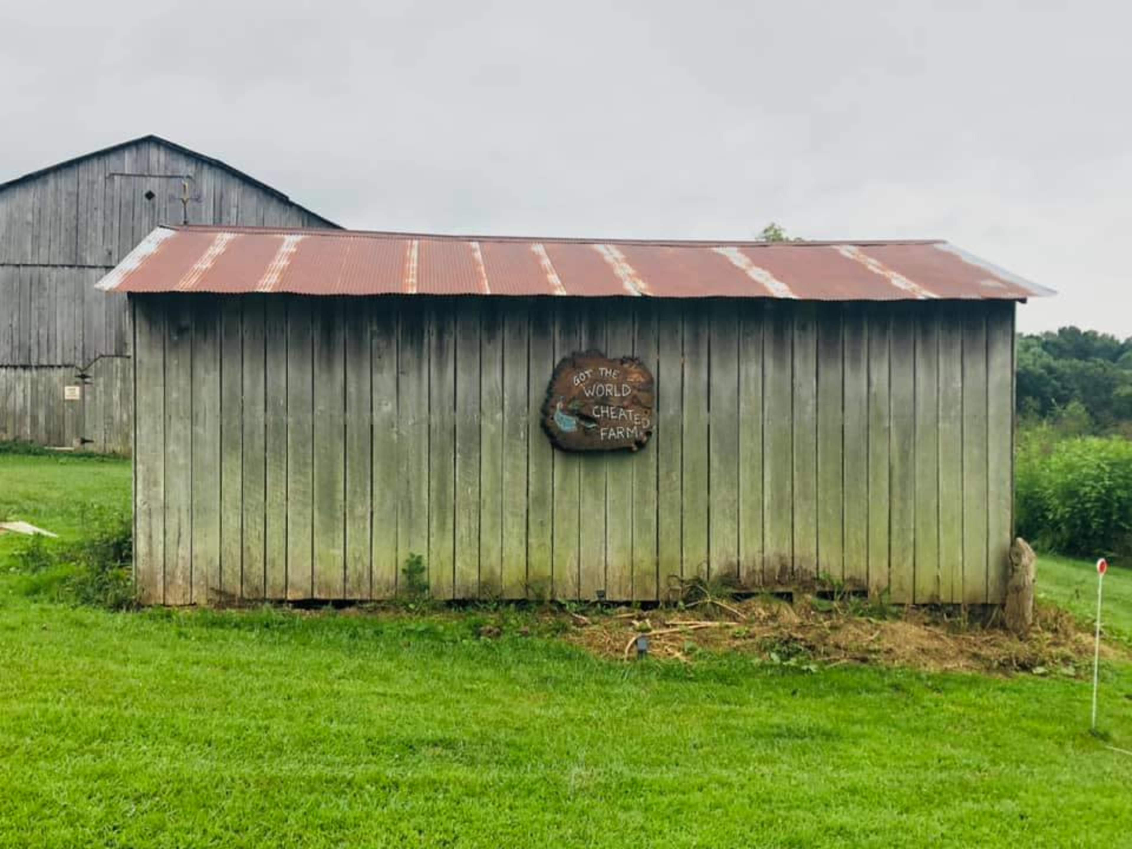 A weathered wooden barn with a tin roof displays a sign reading "World's Largest Shelter Farm" on its side, set against a cloudy sky and grassy field.