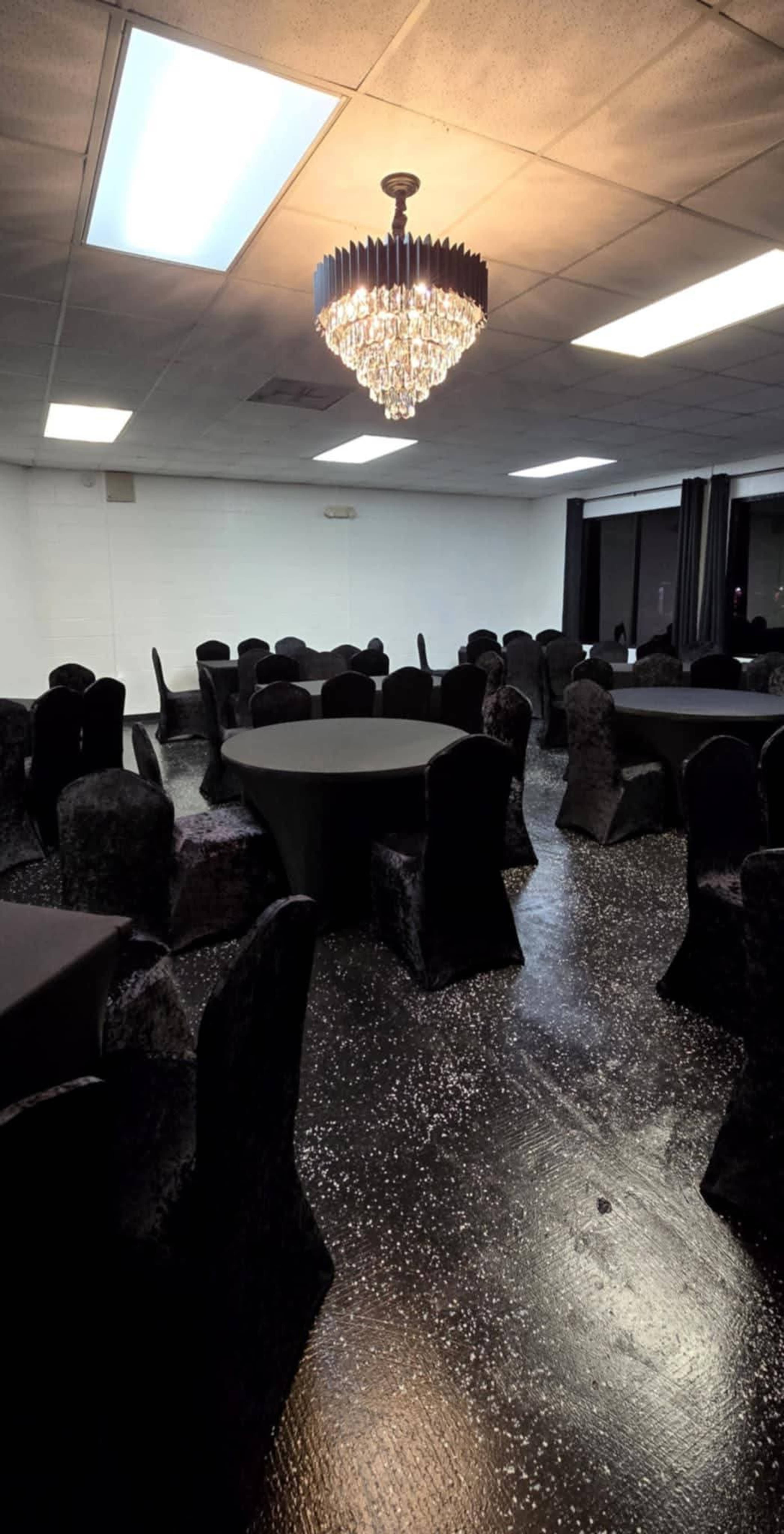 A spacious event room with black chairs and tables, illuminated by a crystal chandelier overhead.