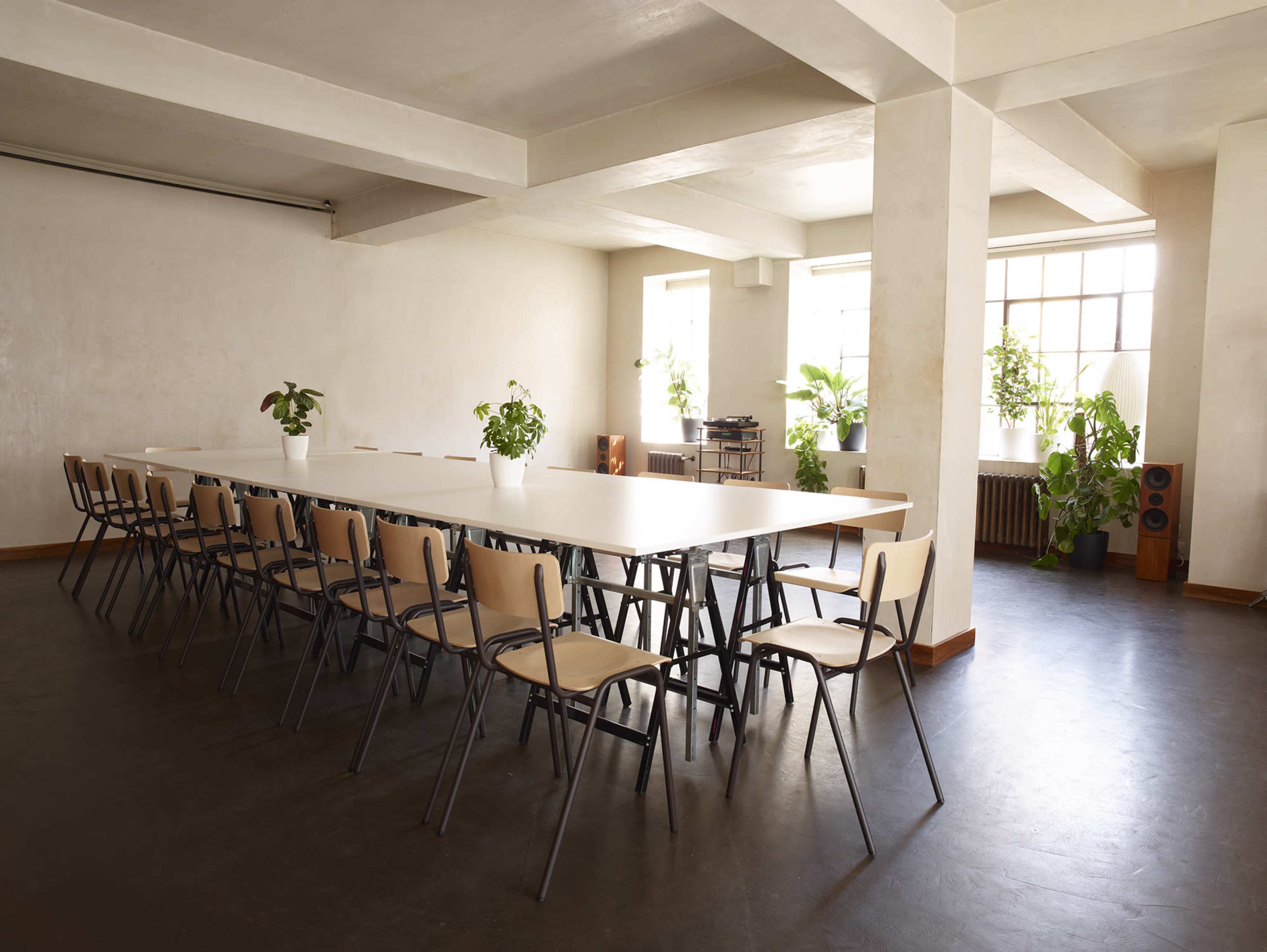 A long, minimalist table with chairs is set in a spacious room filled with plants and large windows.