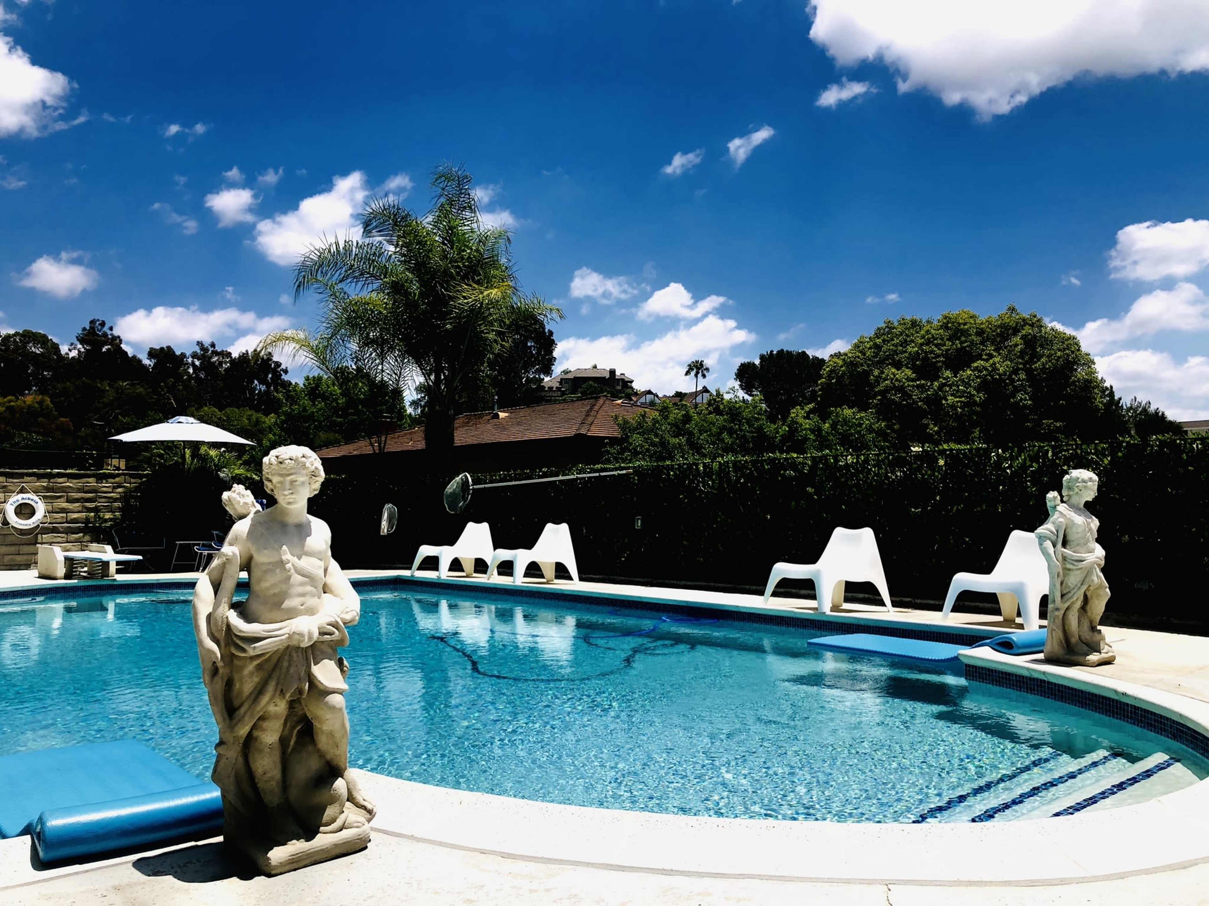 A swimming pool features two white statues and several lounge chairs under a clear blue sky with scattered clouds.