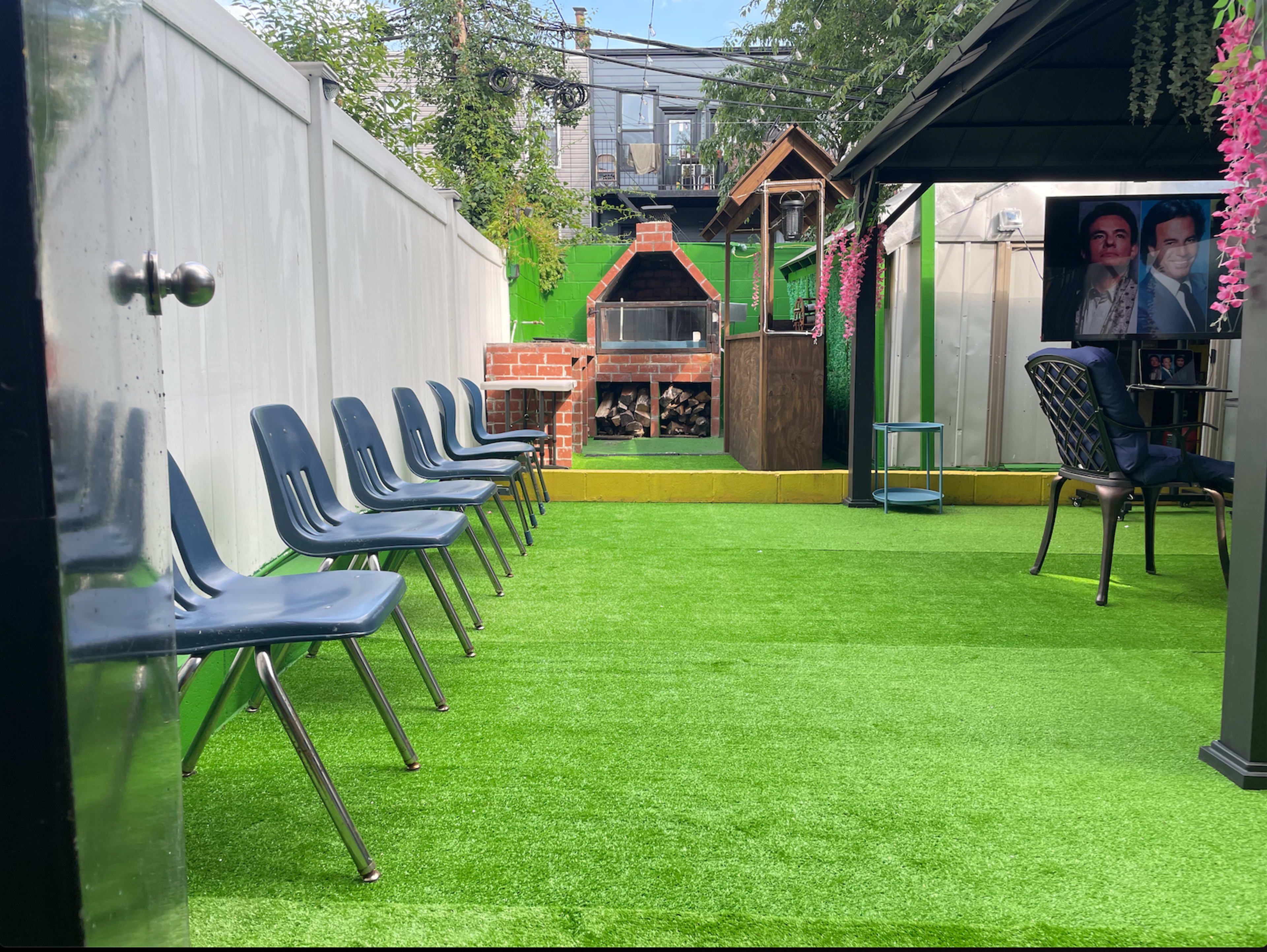 An outdoor space with a row of blue chairs on artificial grass, a barbecue area in the background, and a small gazebo with portraits visible.