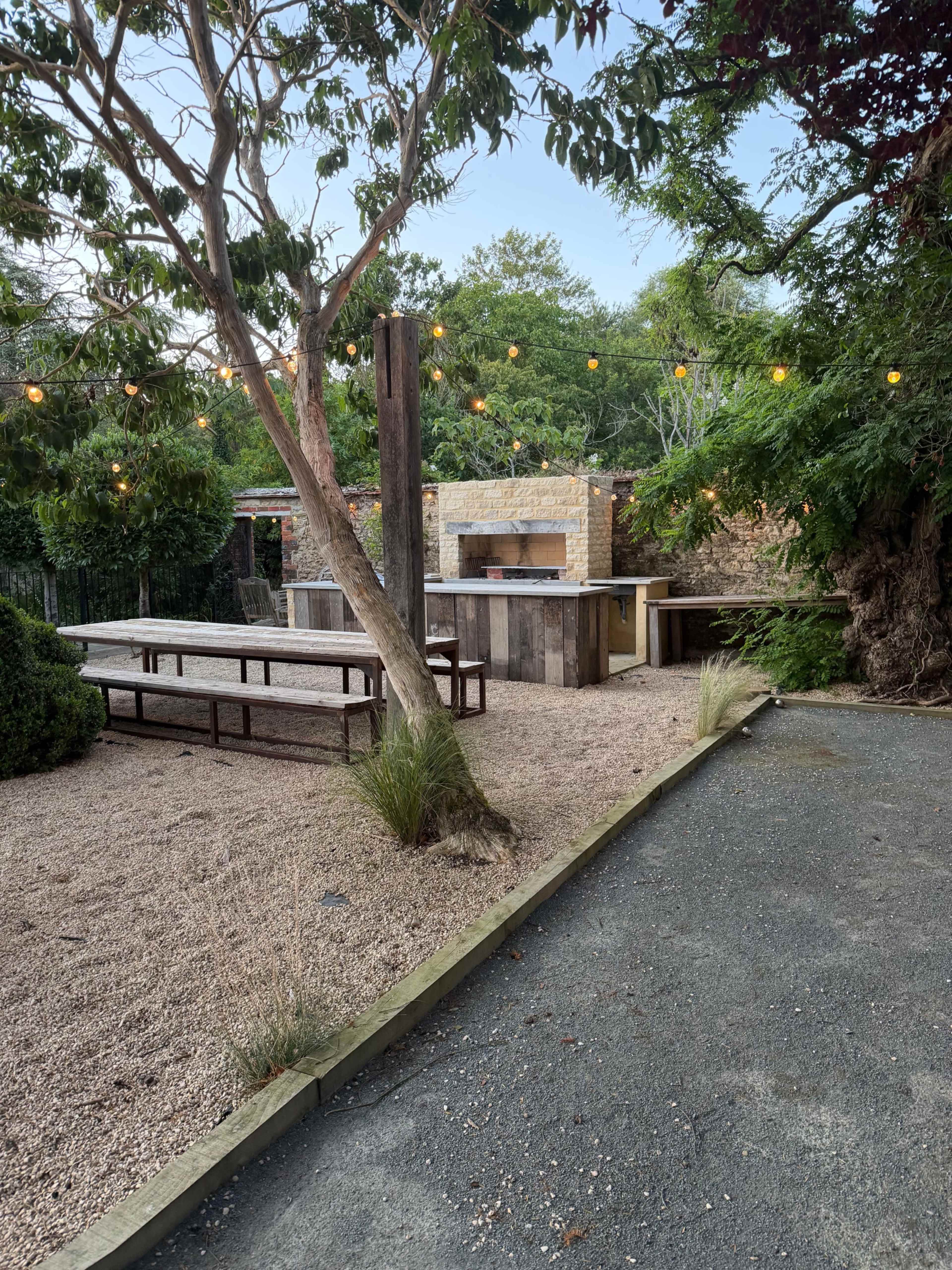 The outdoor space features a gravel area with long wooden tables, a stone fireplace, and string lights overhead, surrounded by greenery and trees.