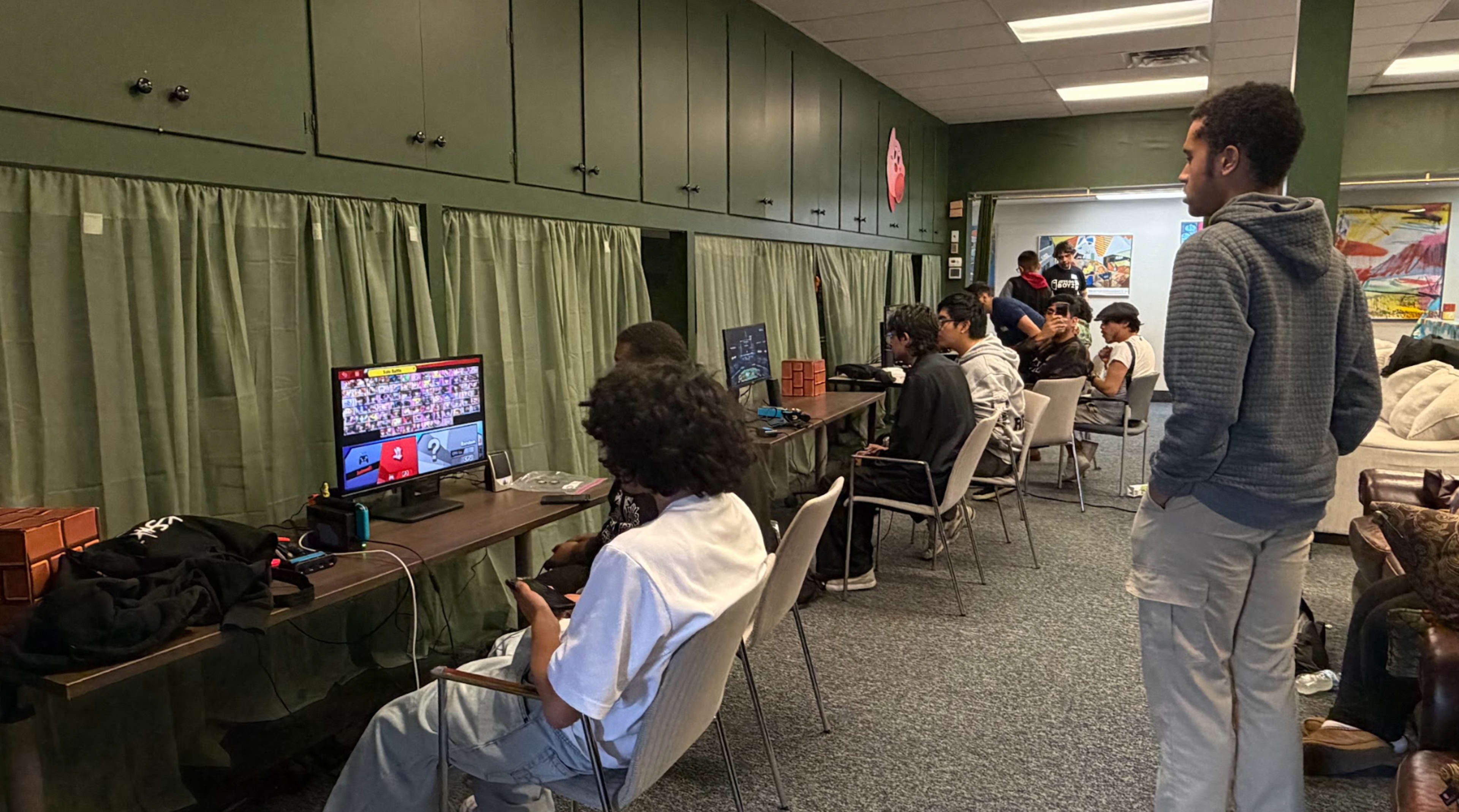 A group of young people are seated at tables in a room playing video games on multiple screens while one person stands and watches.