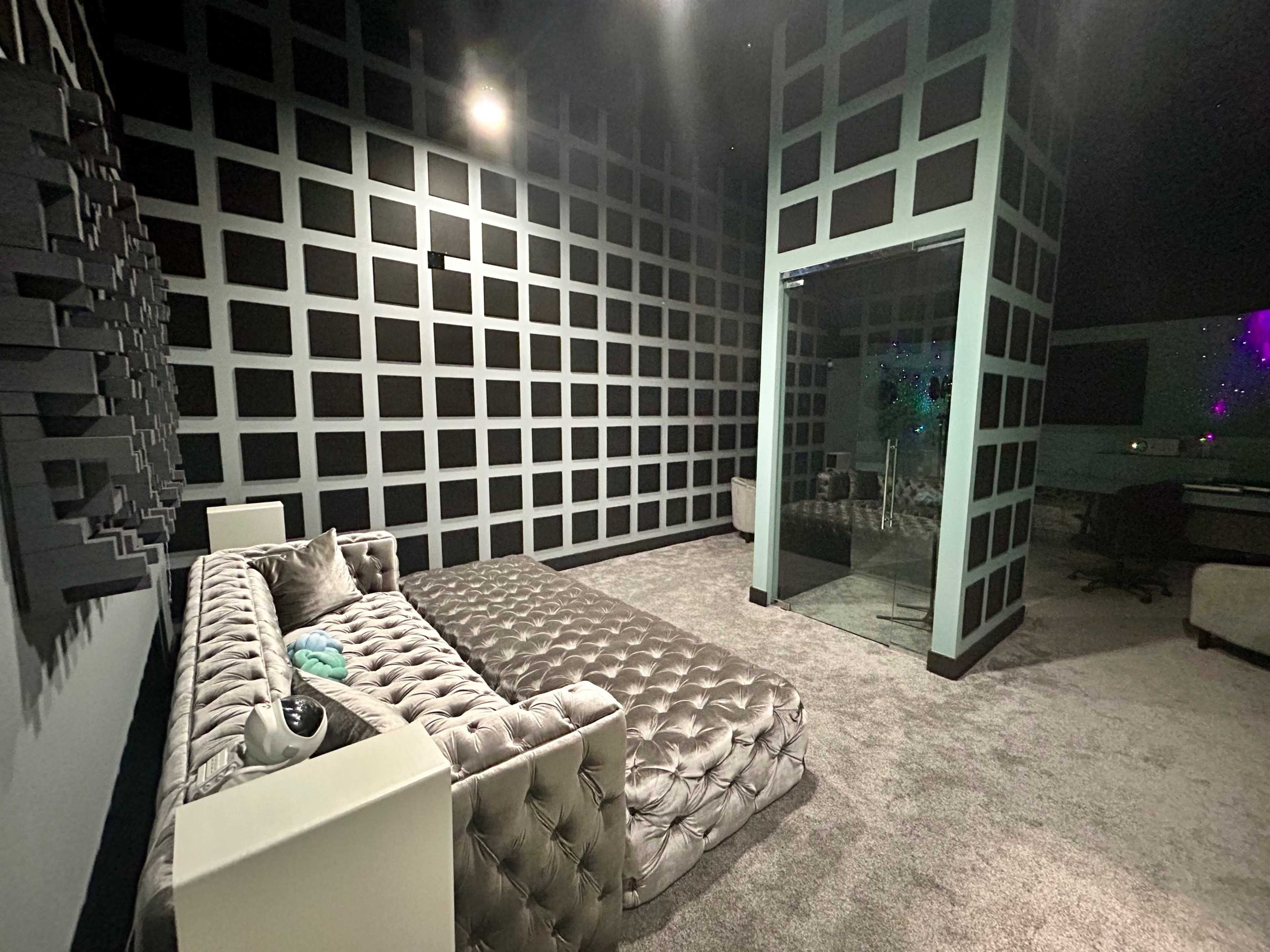 The image shows a modern, stylistically designed room featuring a tufted sofa, a glass-walled corner, and textured wall panels arranged in a grid pattern.