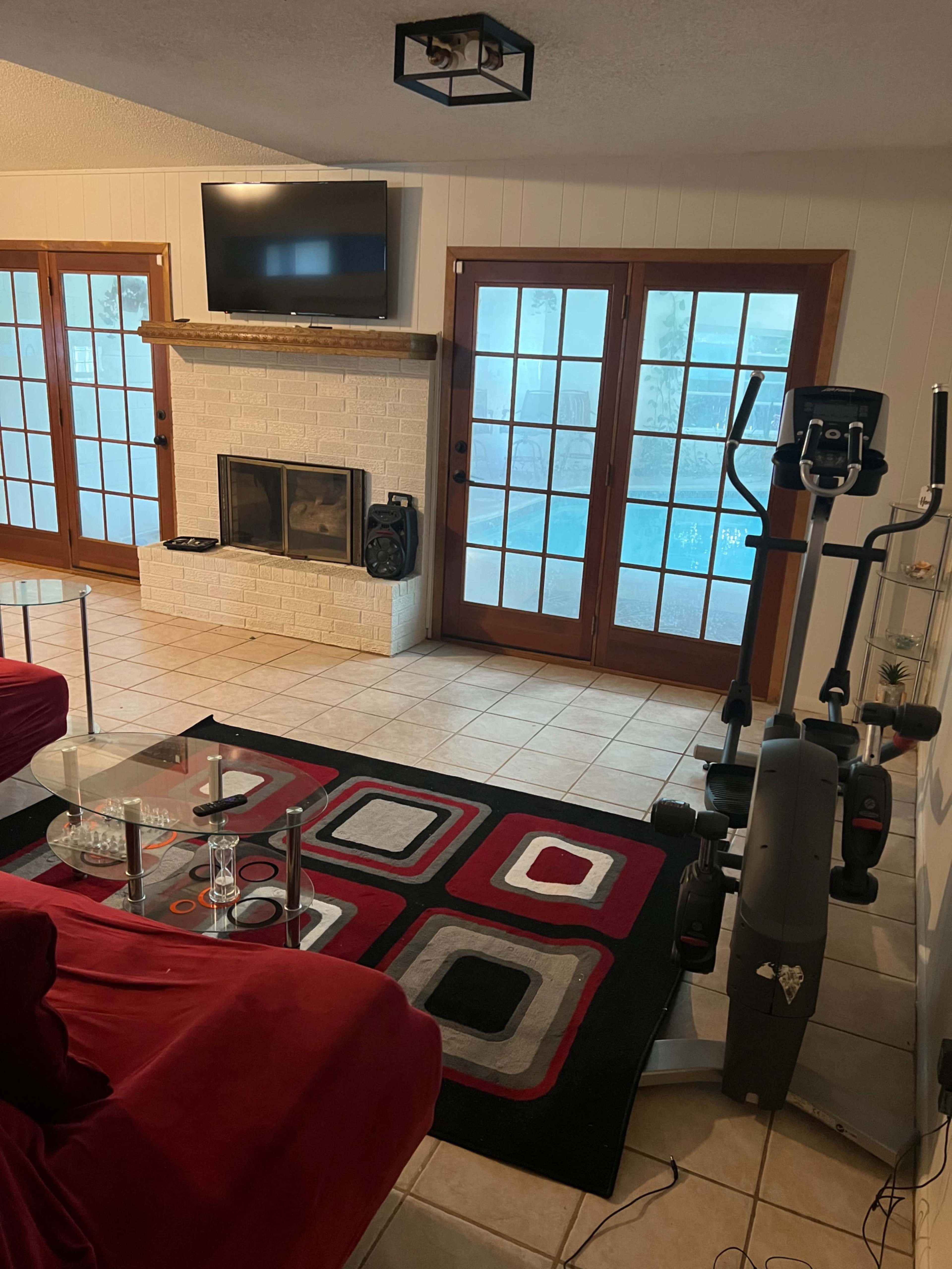 A living room with a red couch, a glass coffee table, a fireplace on a white brick wall, a TV mounted above the mantel, and glass doors leading to a pool area.