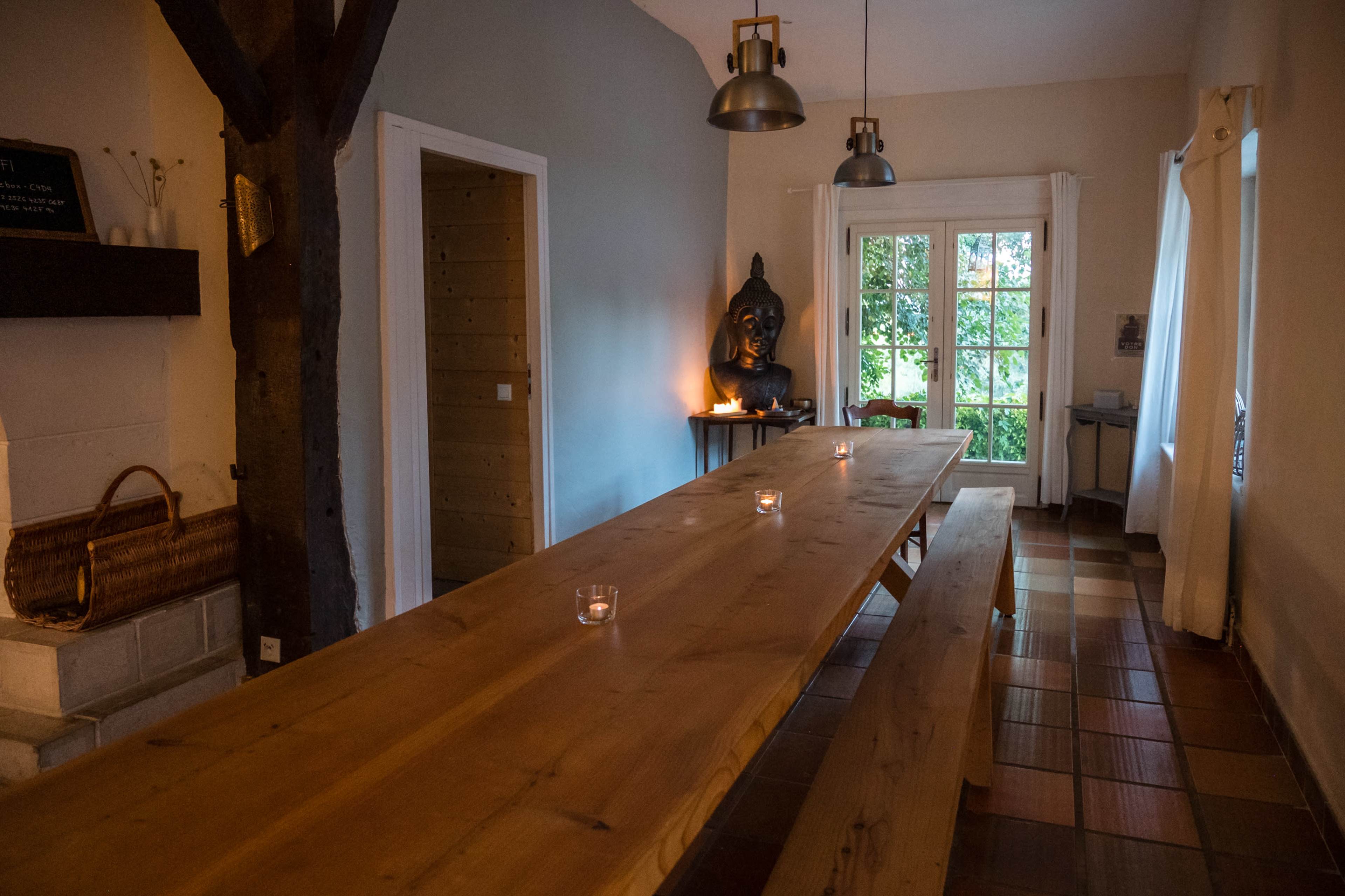 A long wooden table with candles is set in a room featuring large windows and a statue in the corner.